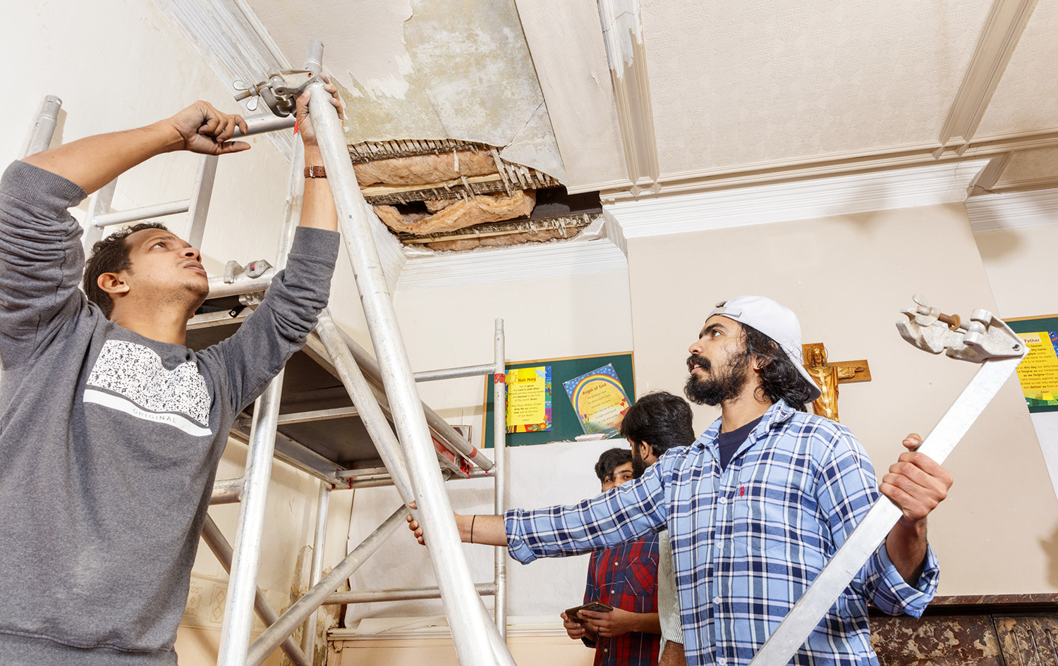 Two men erect a scaffold frame underneath a damaged ceiling.