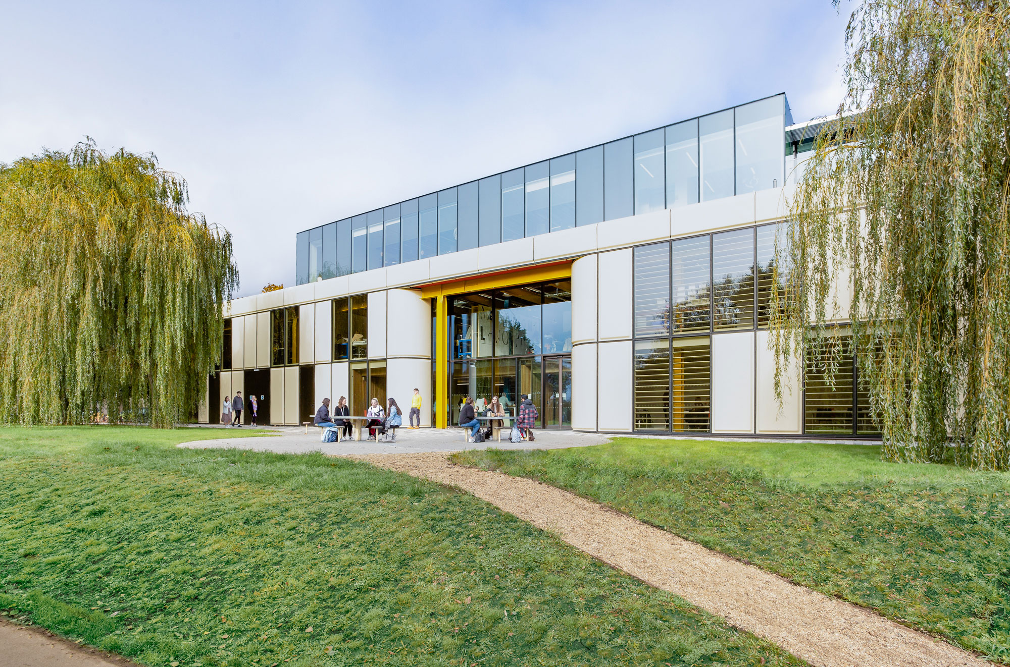 A modern former industrial building with glass and fibreglass panelled elevation, outside are groups of young people sitting at tables.