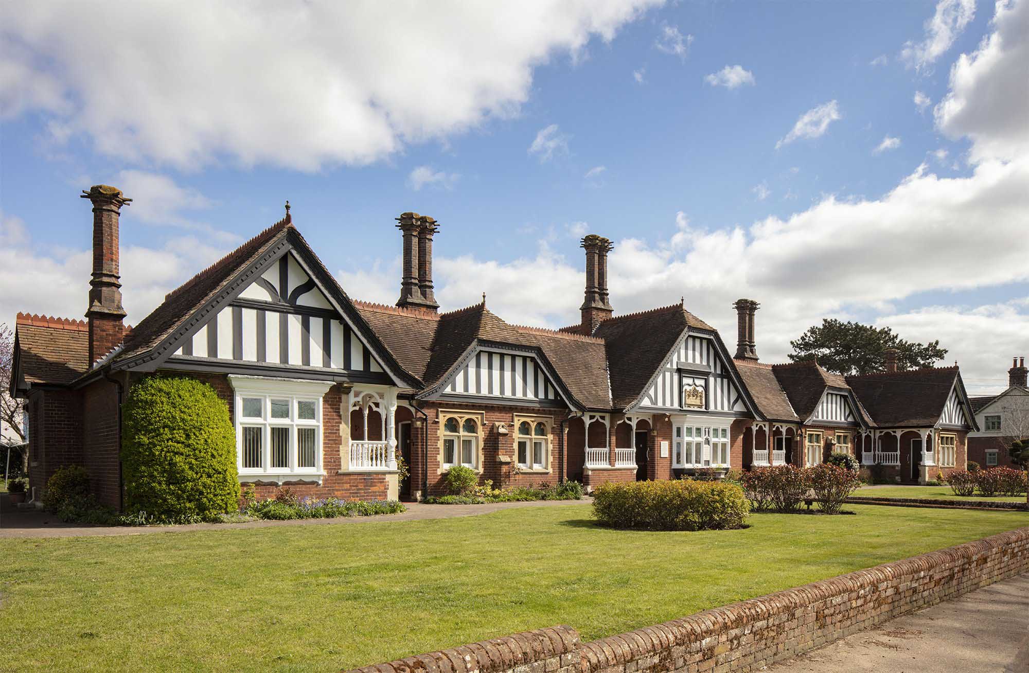 St Edmund's Almshouse showing manicured lawn, short brick wall and the red brick building with gables with half-timbered decoration and decorated chimney stacks.