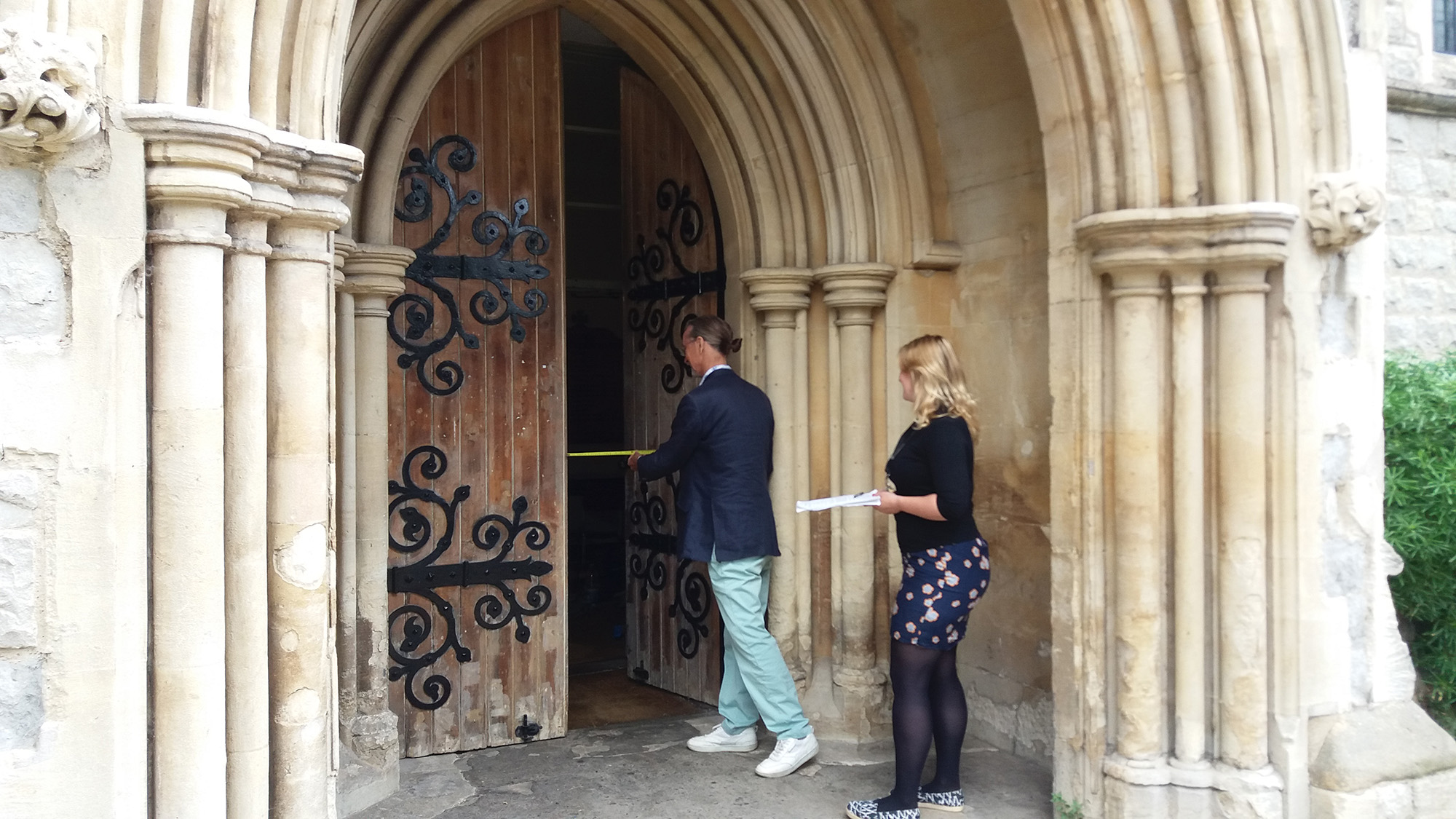 Two people stand to audit the width of the opening of a church entrance. One person is measuring the gap in the open door with a tape measure, while the other holds a clipboard.