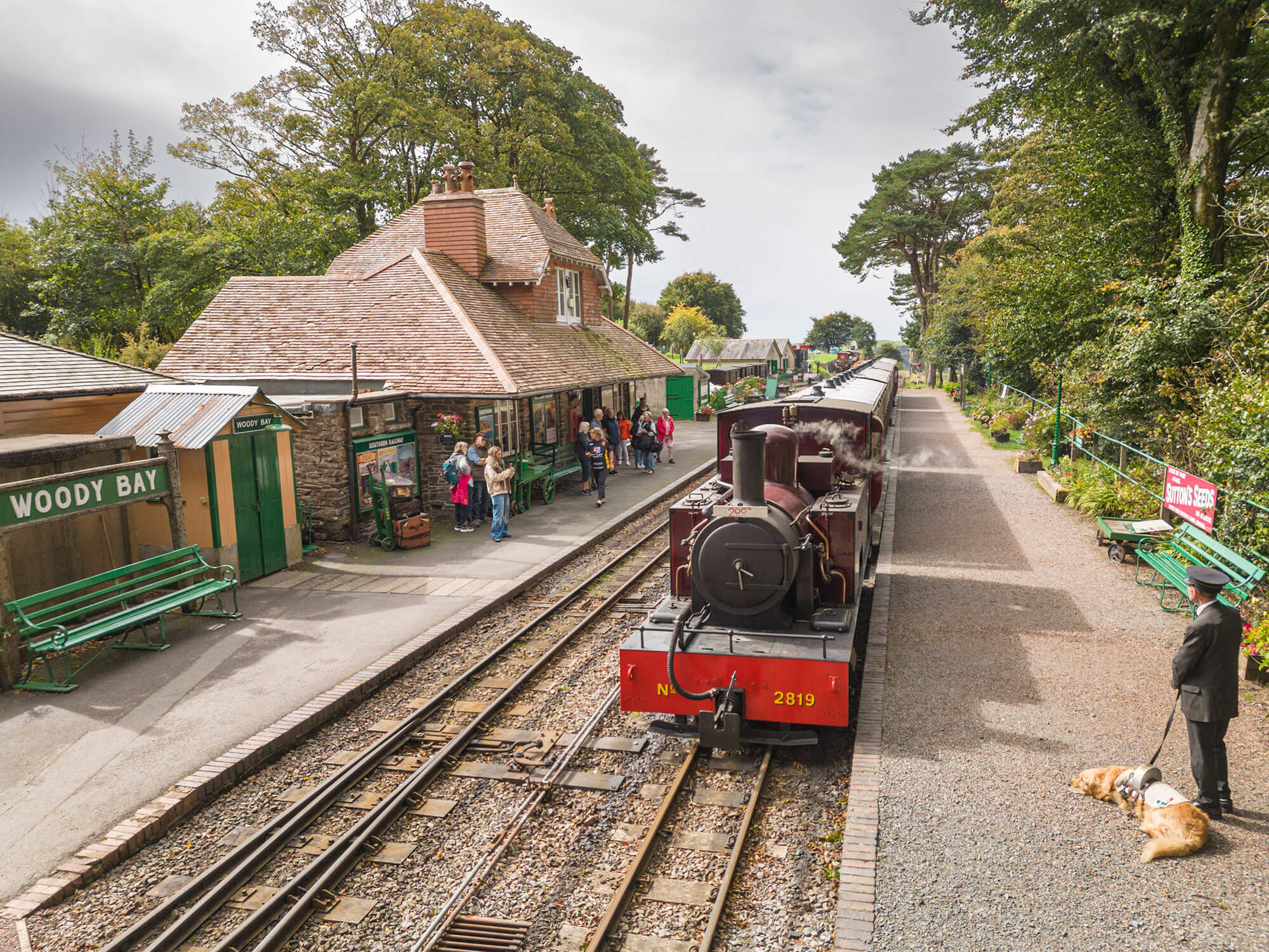 Image of a station with a red steam train pulling in. On the left hand side platform a sign reads Woody Bay next to a small stone building, on the right is a person in a railway uniform with a dog.