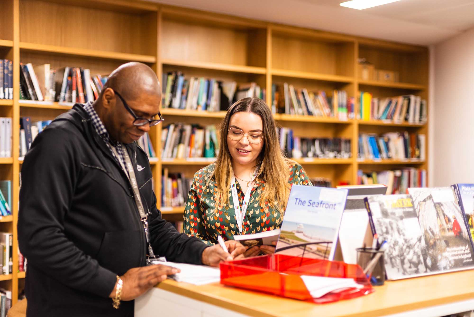 Two staff members stand in discussion, looking down at a piece of paper. In the background are bookshelves. In the foreground, a plastic in-tray and a display of books are arranged along the top of a half-height shelving unit.