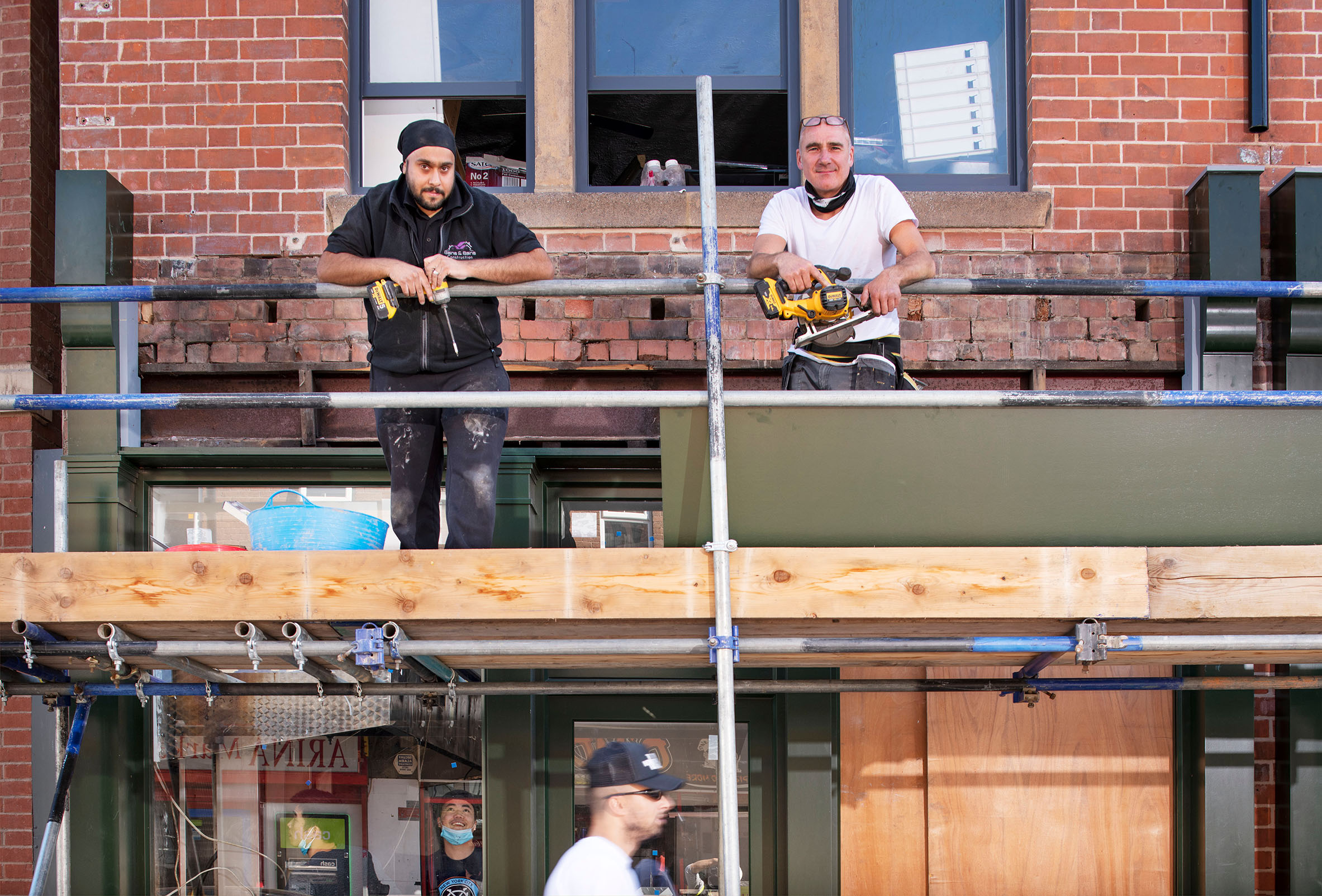 Two men pose for a photo on scaffolding in front of a brick building they're currently restoring.