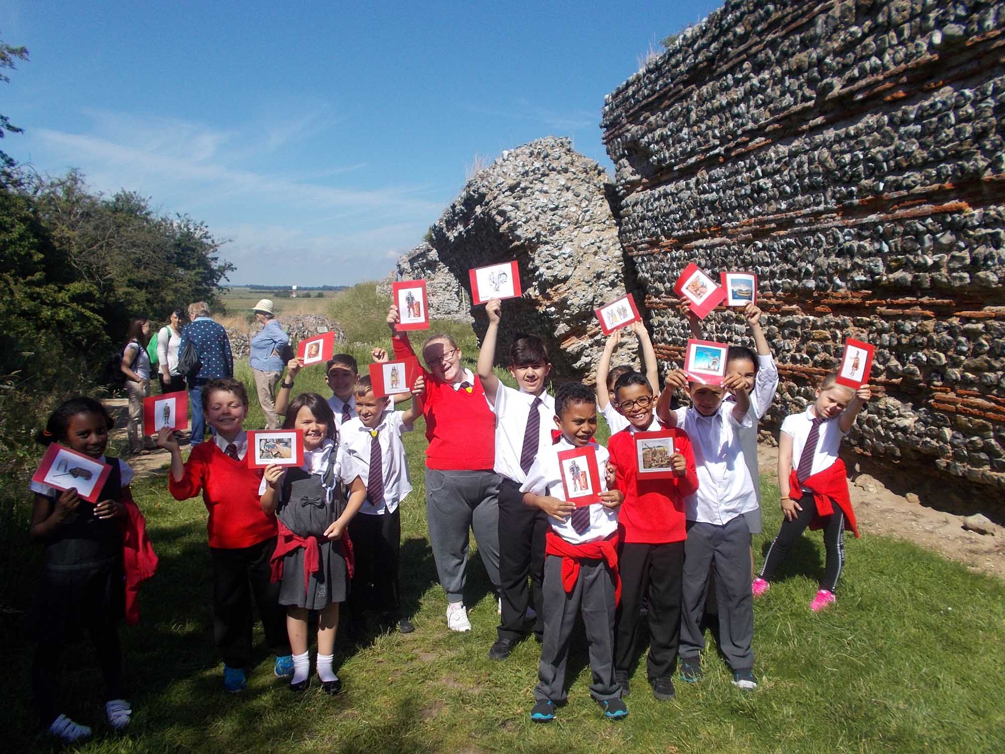 Primary school children in uniform, on a sunny day, hold aloft creative pictures next to a historic Great Yarmouth town wall.