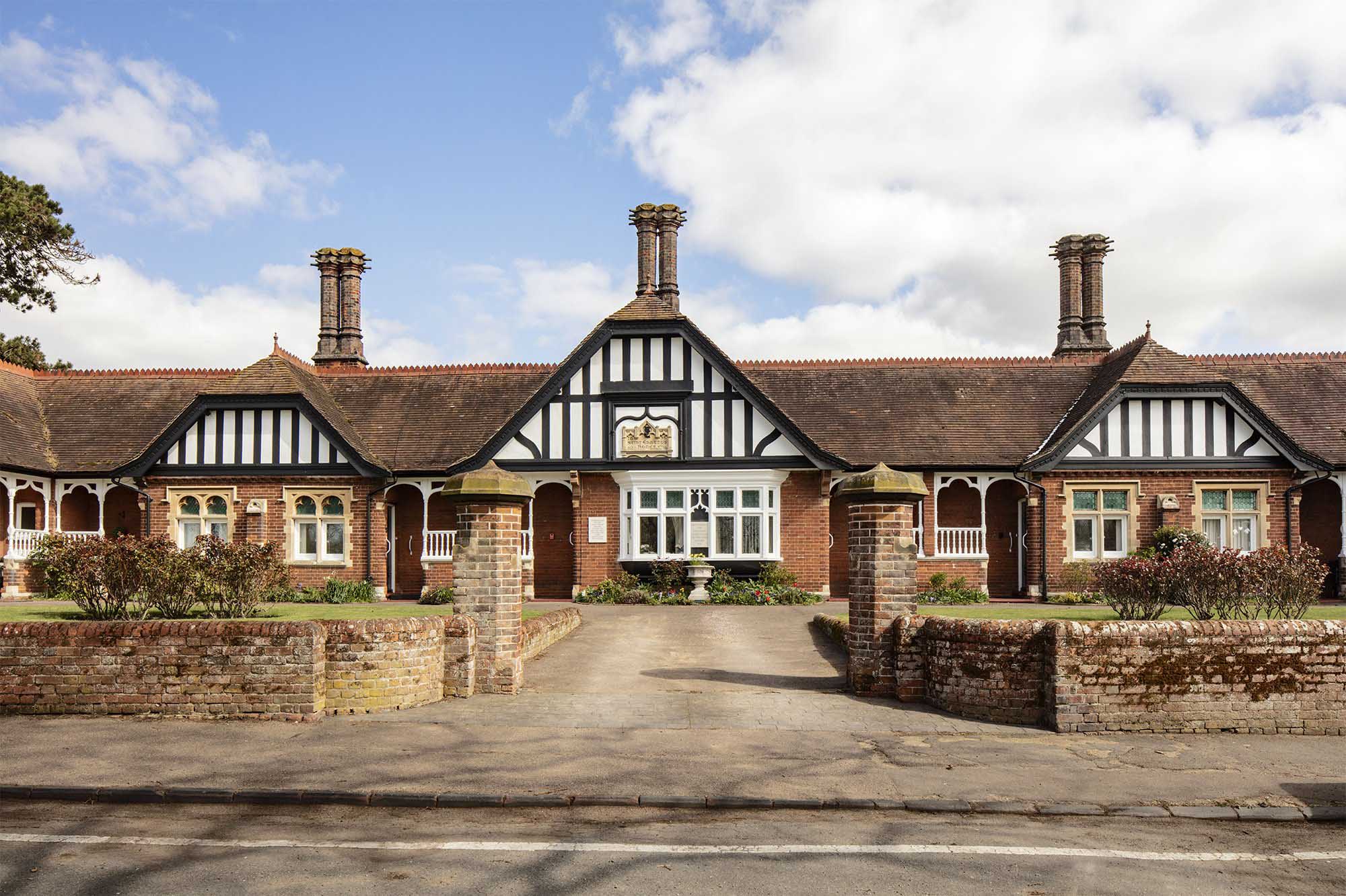 A low-level brick building with three gables with half-timbered decoration in black and white and pairs of star-topped chimney stacks.
