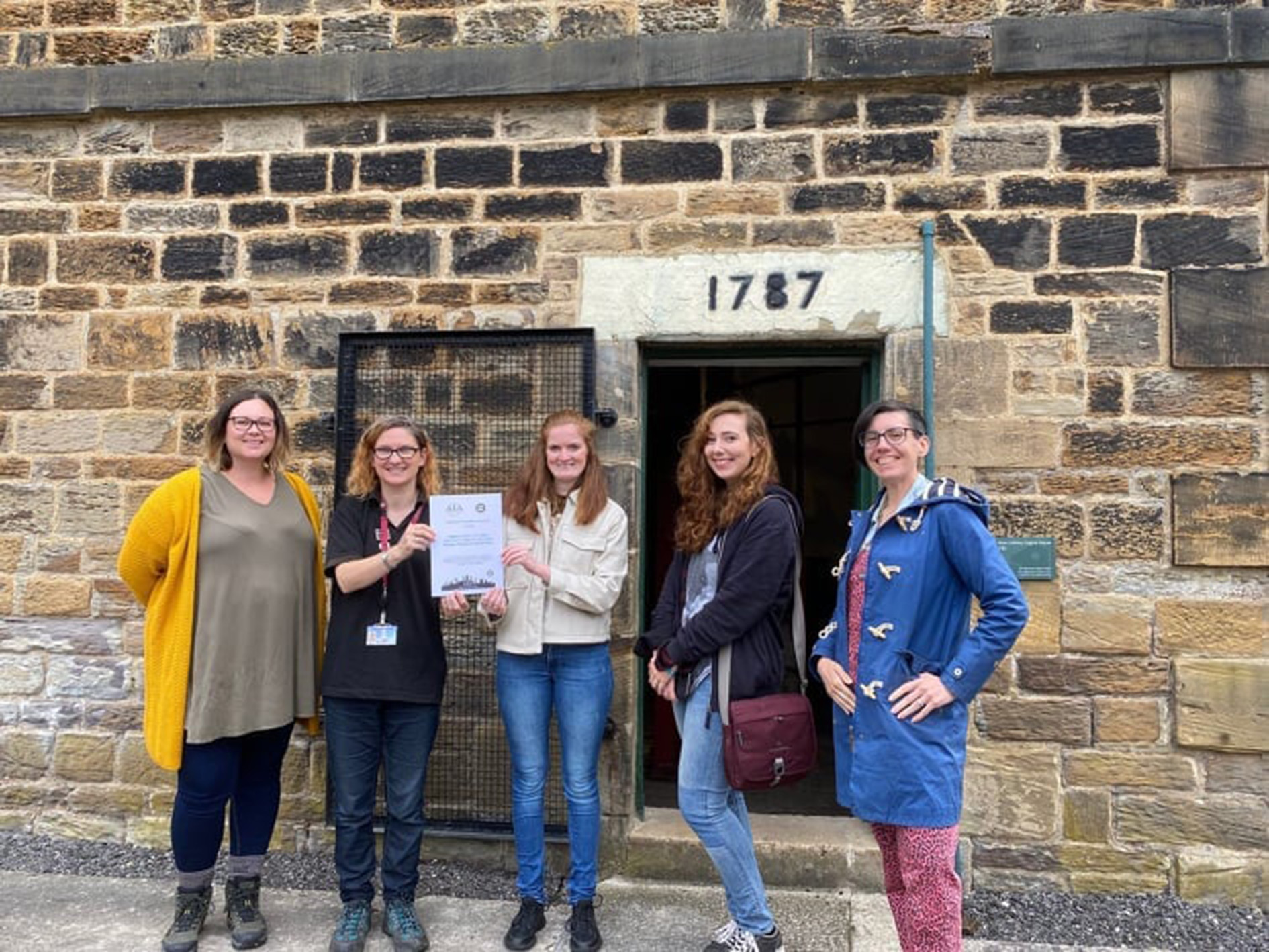 A group of five standing in front of a stone building with a sign that reads "1787." Two in the centre hold a certificate. The group is smiling and dressed casually in a mix of jackets, cardigans, and jeans.