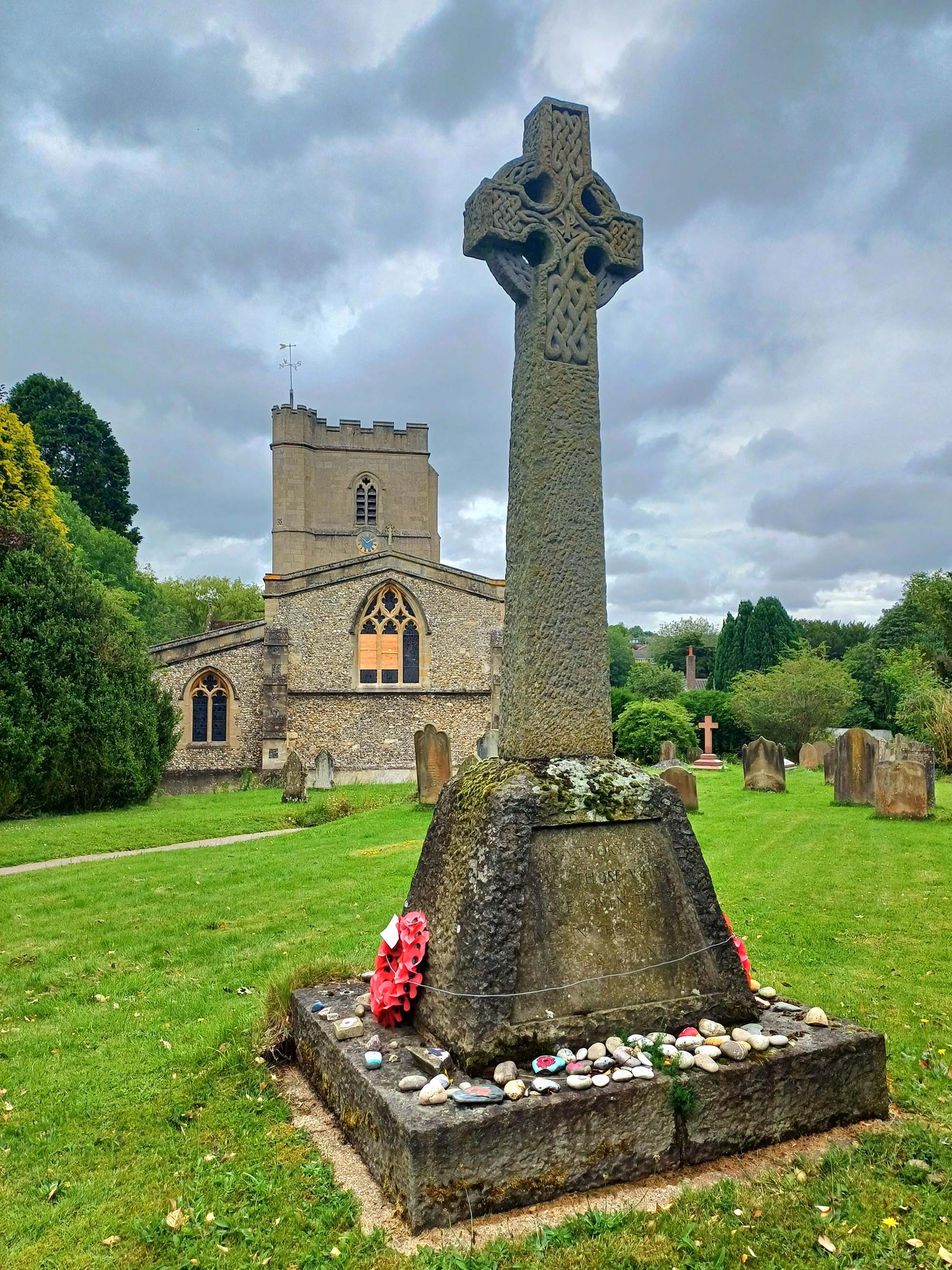 A Celtic cross with celtic decoration, stands on a plinth in a churchyard. The church tower can be seen in the background.