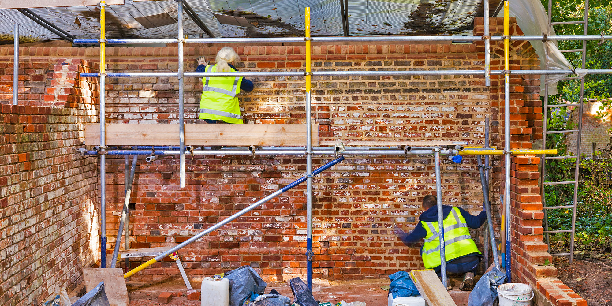 A pair of workers in PPE repoint a landscape wall from a scaffold. 
