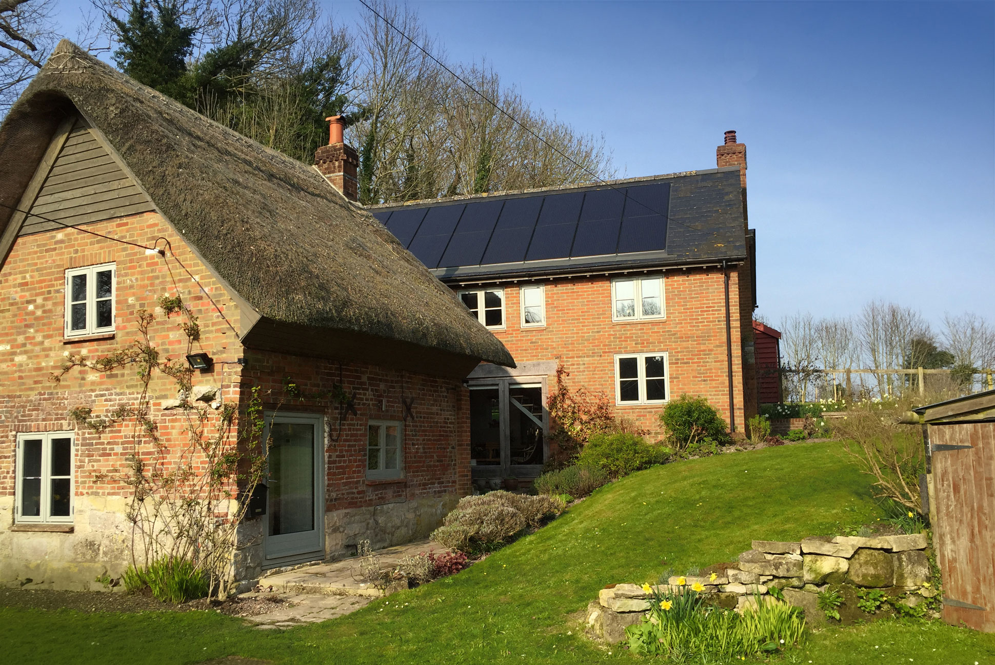 A photo of two detatched domestic buildings perpendicular to one another. The one in the foreground is brick with a thatched roof and the building at the rear is brick with a slate roof. The rear building's roof has a row of 8 PV panels.