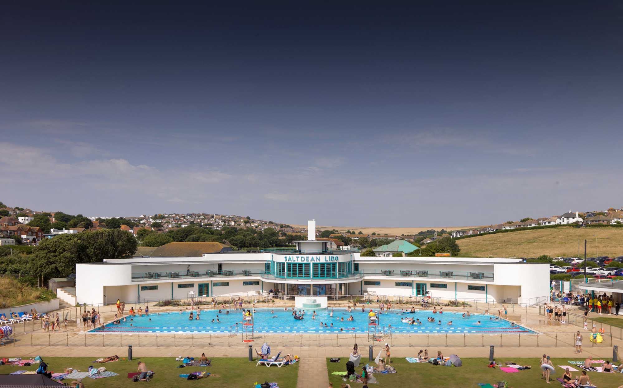 Image of the front of a large art deco building with white walls and blue framed windows. A large blue sign says Saltdean Lido. The lido is visible in the foreground and there are lots of people swimming and sunbathing around the pool.