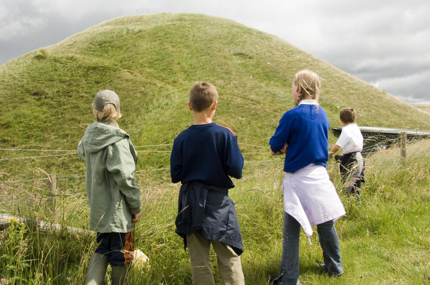A group of children with their backs to camera looking at a neolithic earthwork mound.
