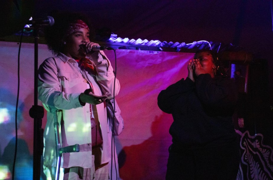 Two people stand on a stage in a room with low lighting. One person is speaking into a microphone while the other cheers them on.