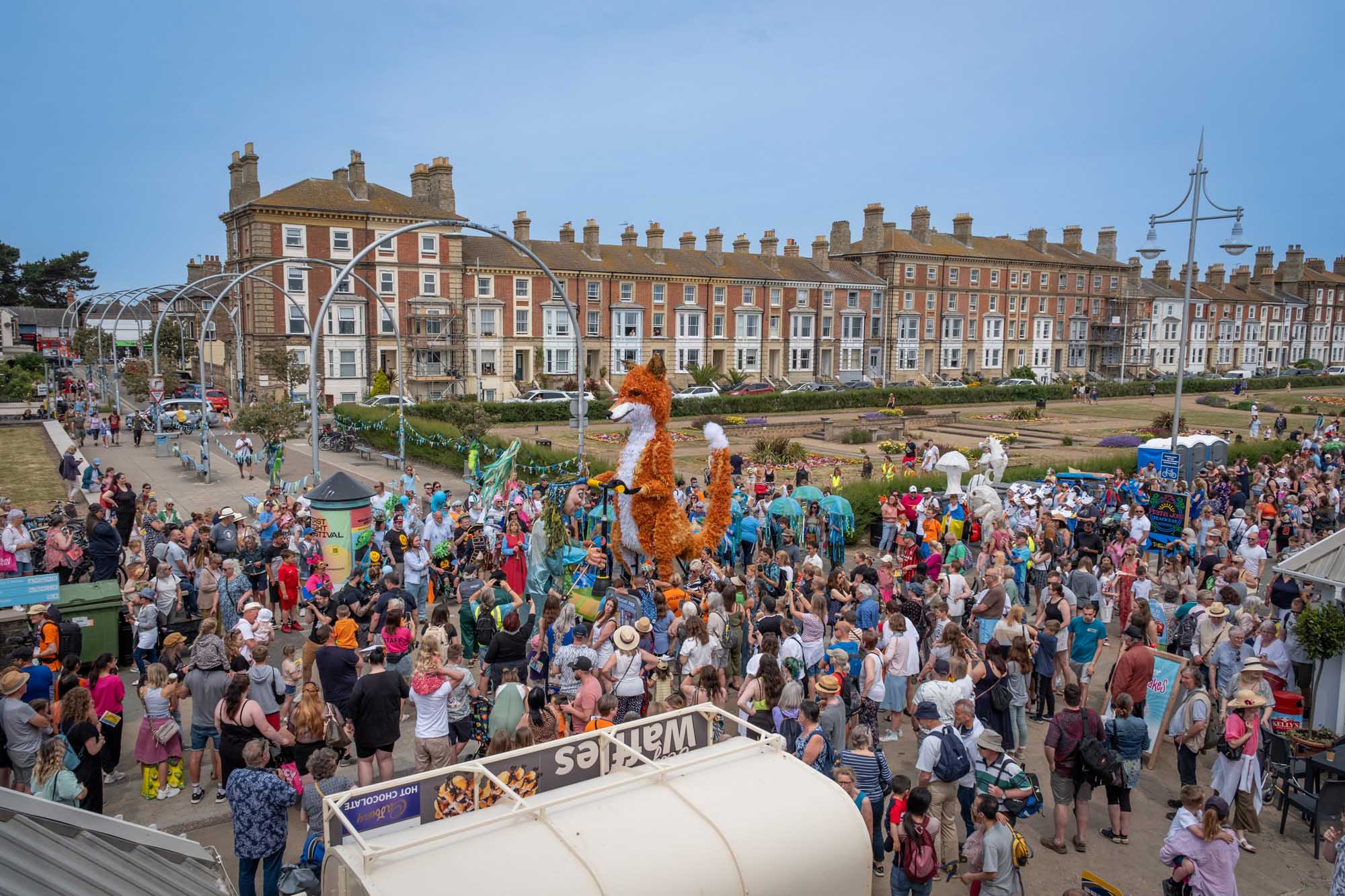 A carnival scene with a crowd, stalls and a fox puppet in the foreground, and a row of buildings in the background. 