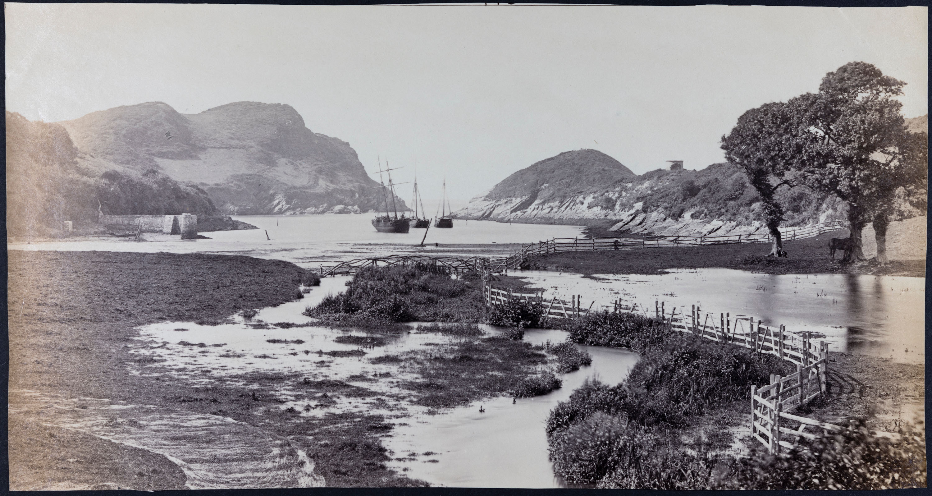 A black and white scenic photograph with boats on a body of water in the distance, flanked by hillside