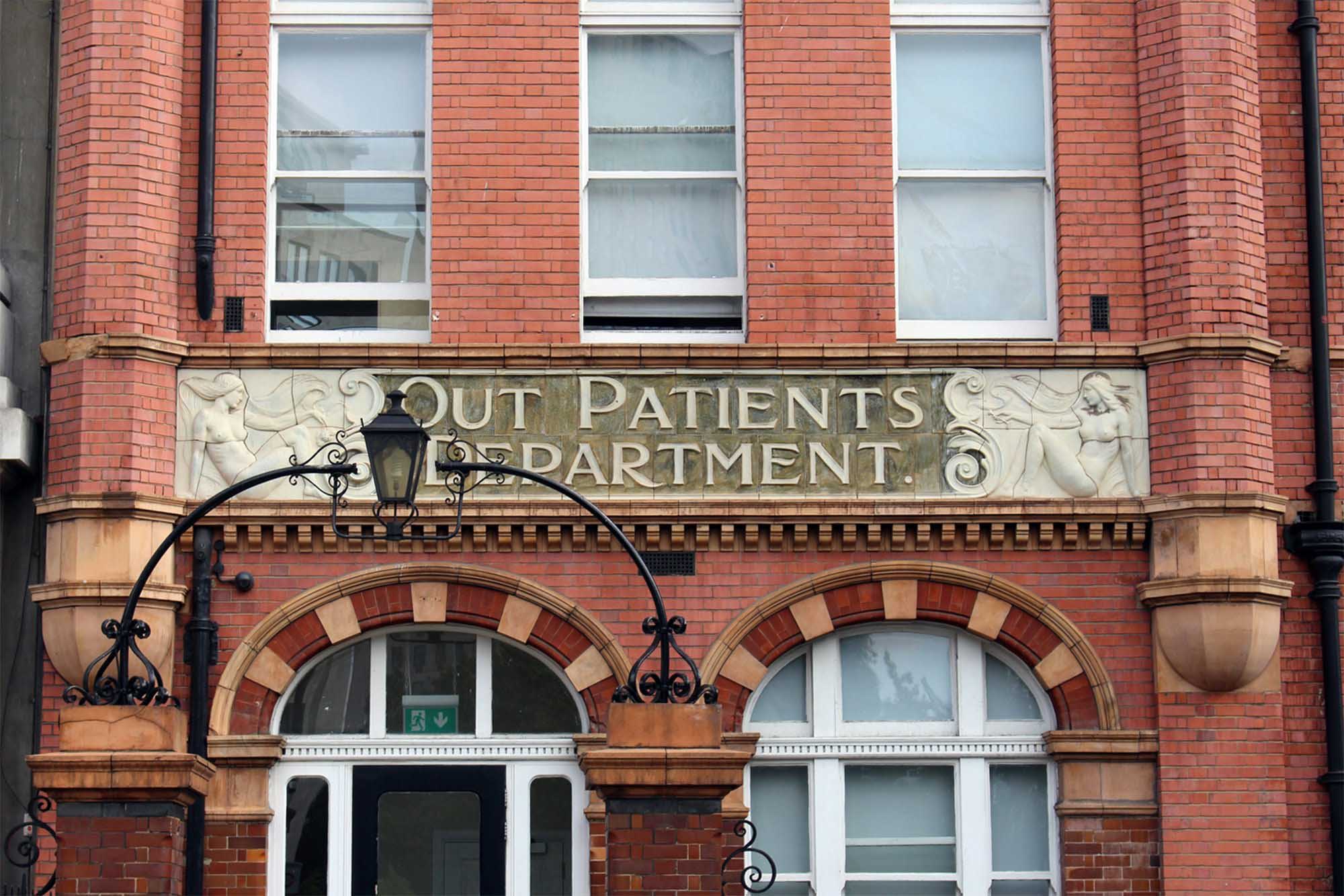 Entrance with the words 'Out Patients Department' engraved in the stone flanked by two women on either side. A lantern above the entrance gate partly obscures the view of the stone carving on the left.