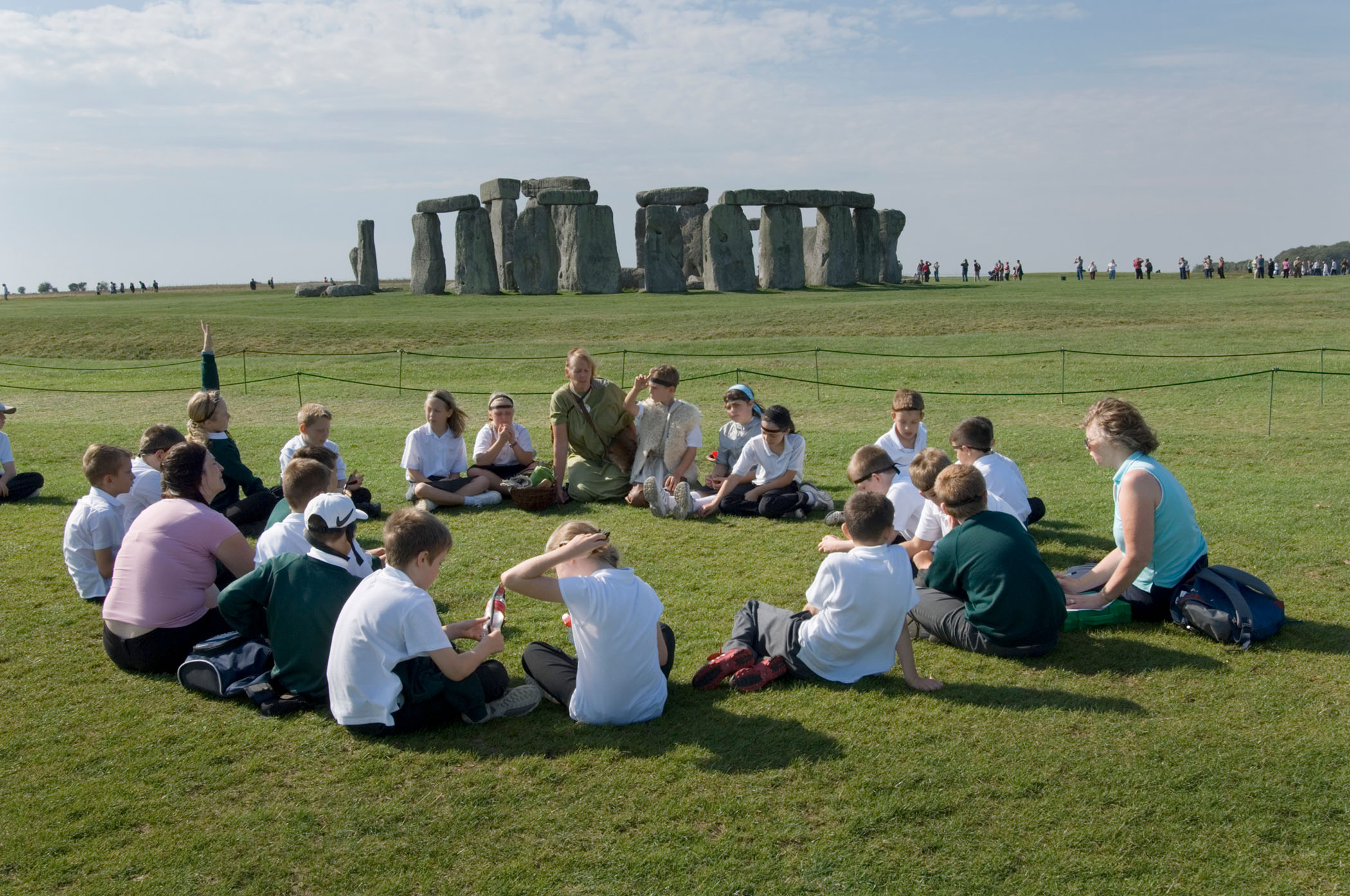A group of students and adults sat in a circle on grass in the foreground, with Stonehenge in the background.