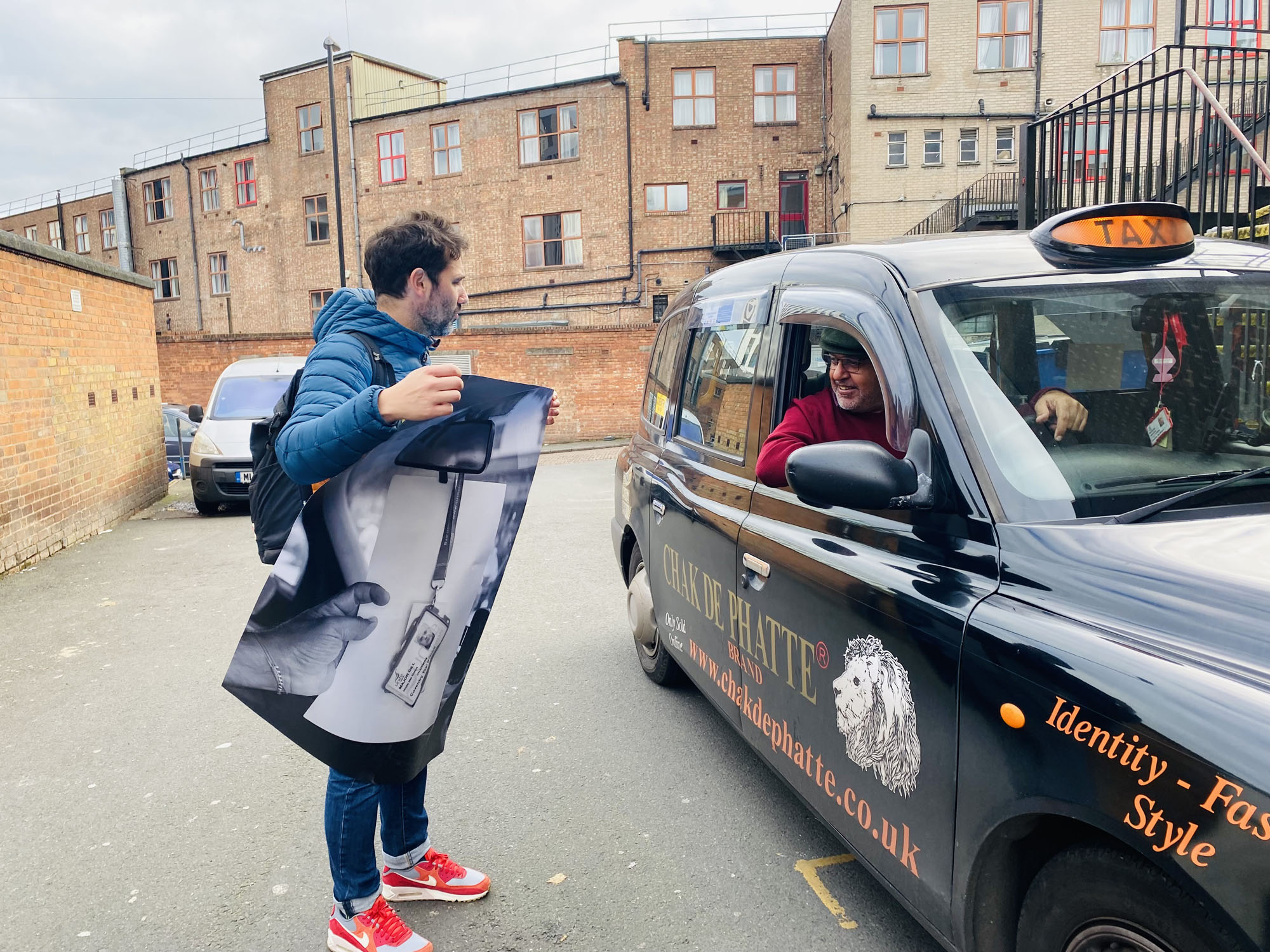 Tim Mills stands with camera equipment, chatting to a taxi driver sitting in his car.