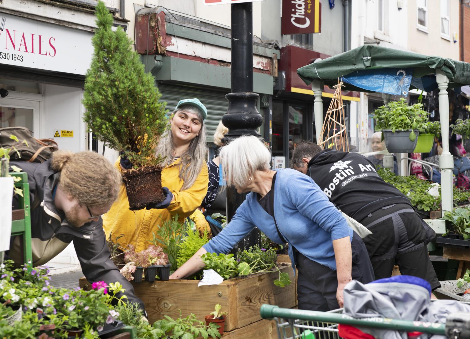 A group of people in a street scene planting raised flower beds. Shop buildings are visible in the background.