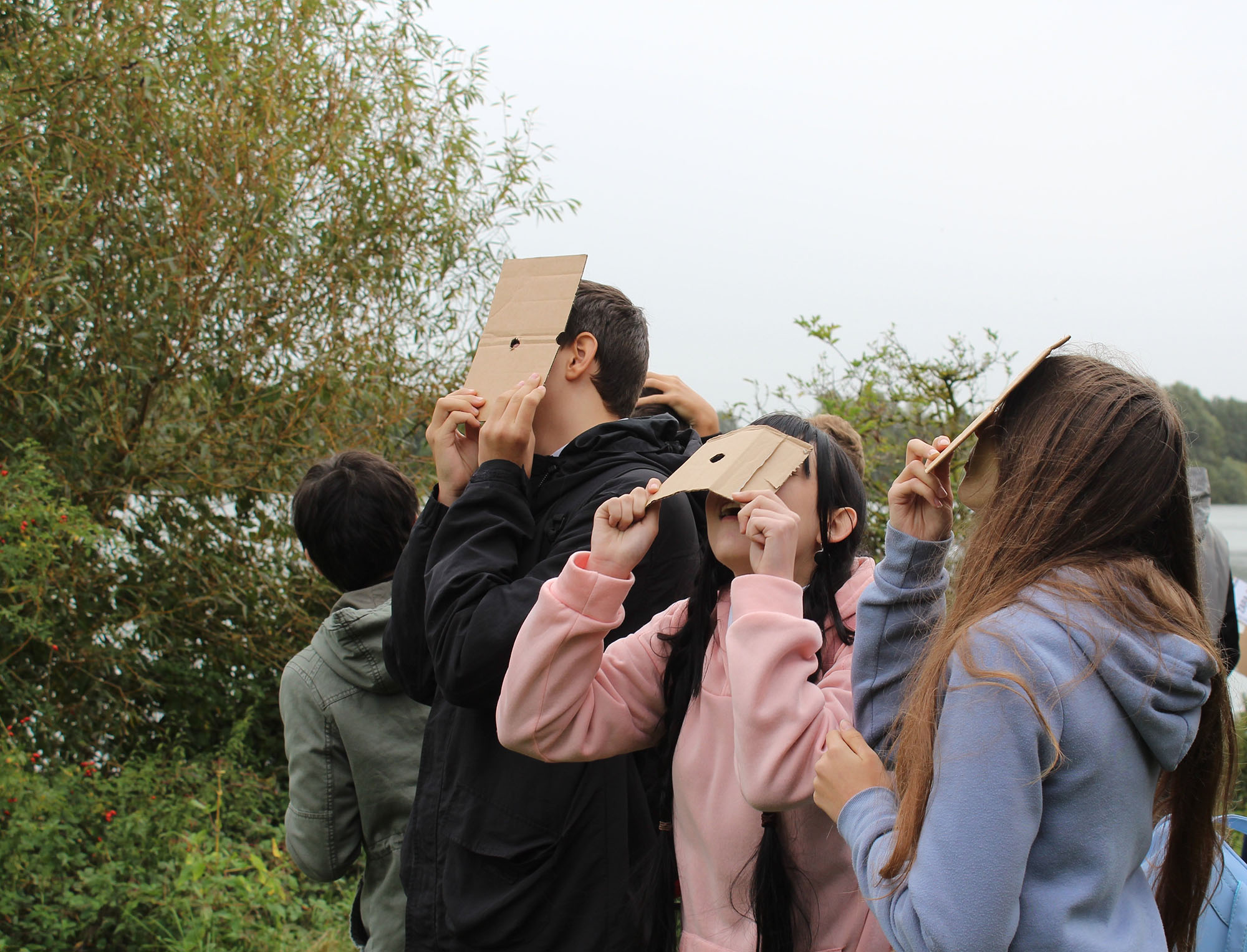 Four young people use pieces of cardboard with a hole in the middle to view the sky. A lake and trees are in the background.