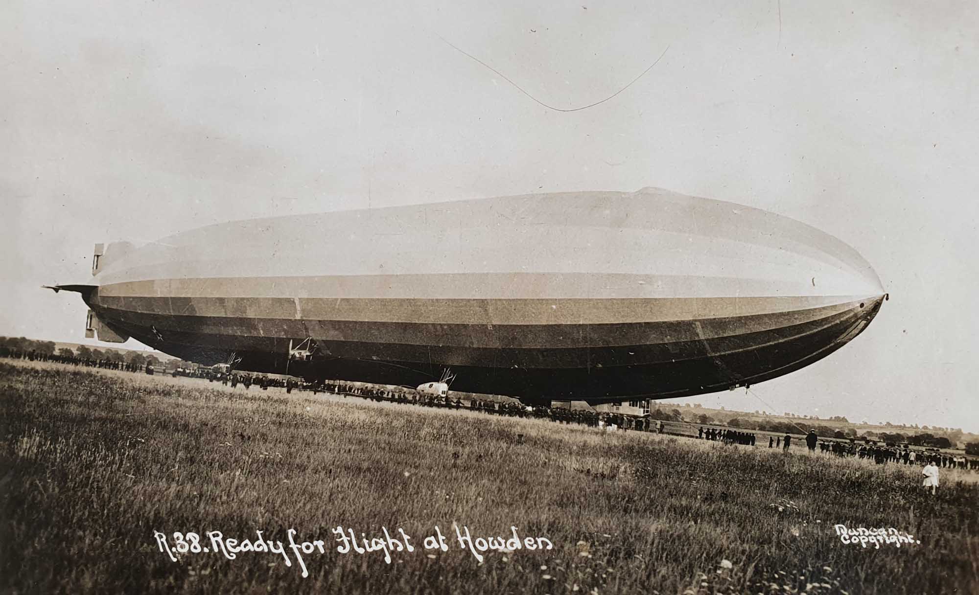 Black and white photo of Airship R.38/ZR 2 preparing for flight. The foreground is flat grassland. The airship sits on the horizon which has a line of dark specks along its entire length. Looking closer, the specks are people, some holding ropes. Hand written text on the bottom edge reads: "R.38. Ready for flight at Howden"