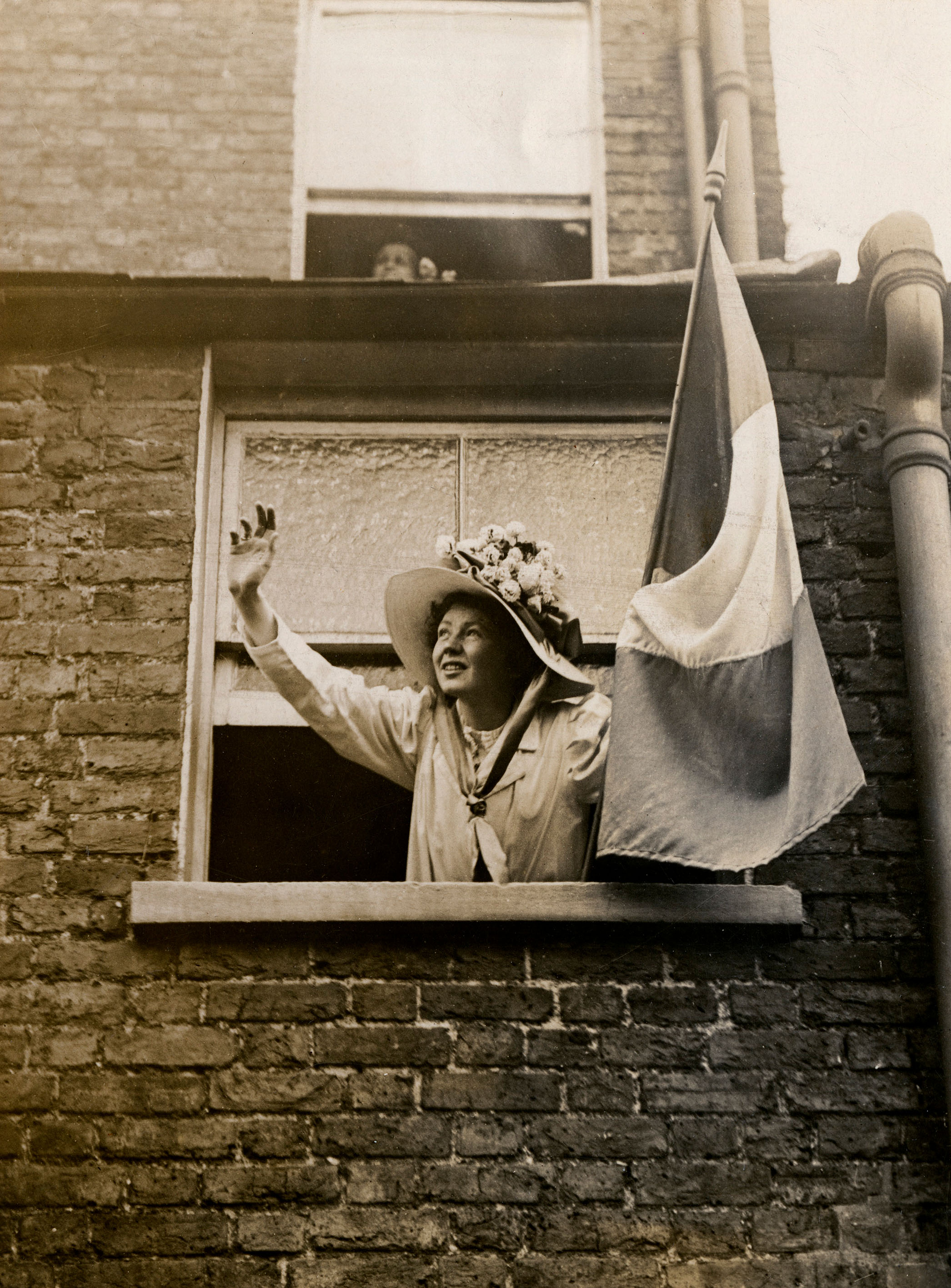Christabel Pankhurst waving out of a window, with a flag in her left hand, wearing large straw hat with flowers on top.