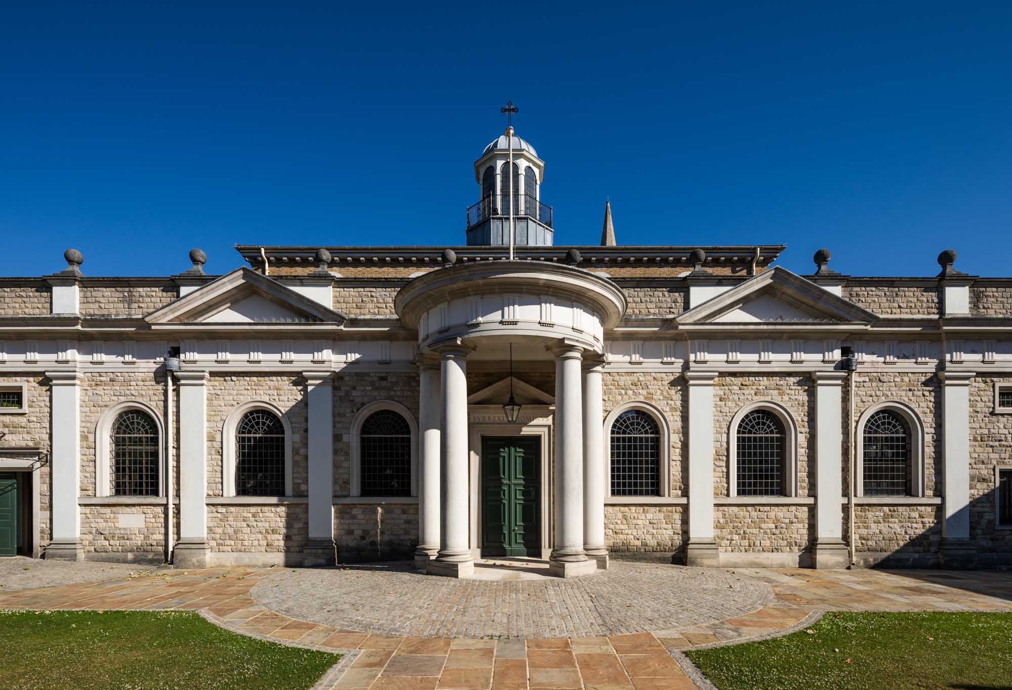 The exterior of Brentwood Cathedral shows a cobbled and paved circular external flooring. A rounded porch with pillars and a long line exterior wall with semi-circular leaded windows and two arches, with a decorative hexagonal tower.