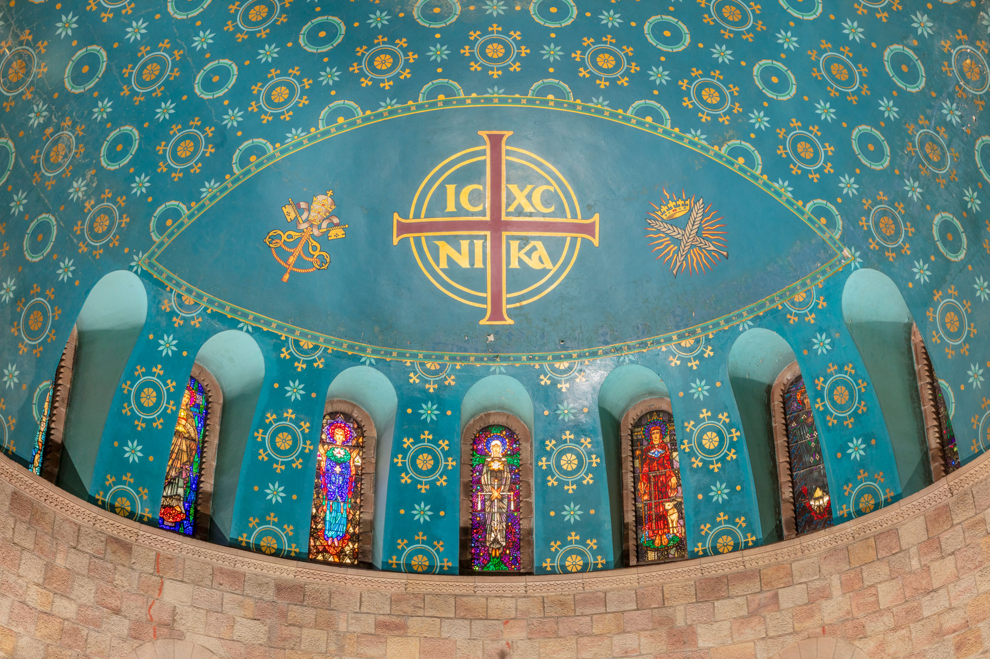 View of a church interior, looking up above to the first floor where 7 stained glass windows in arched recesses form a crescent, set in a dome painted in turquoise and gold.