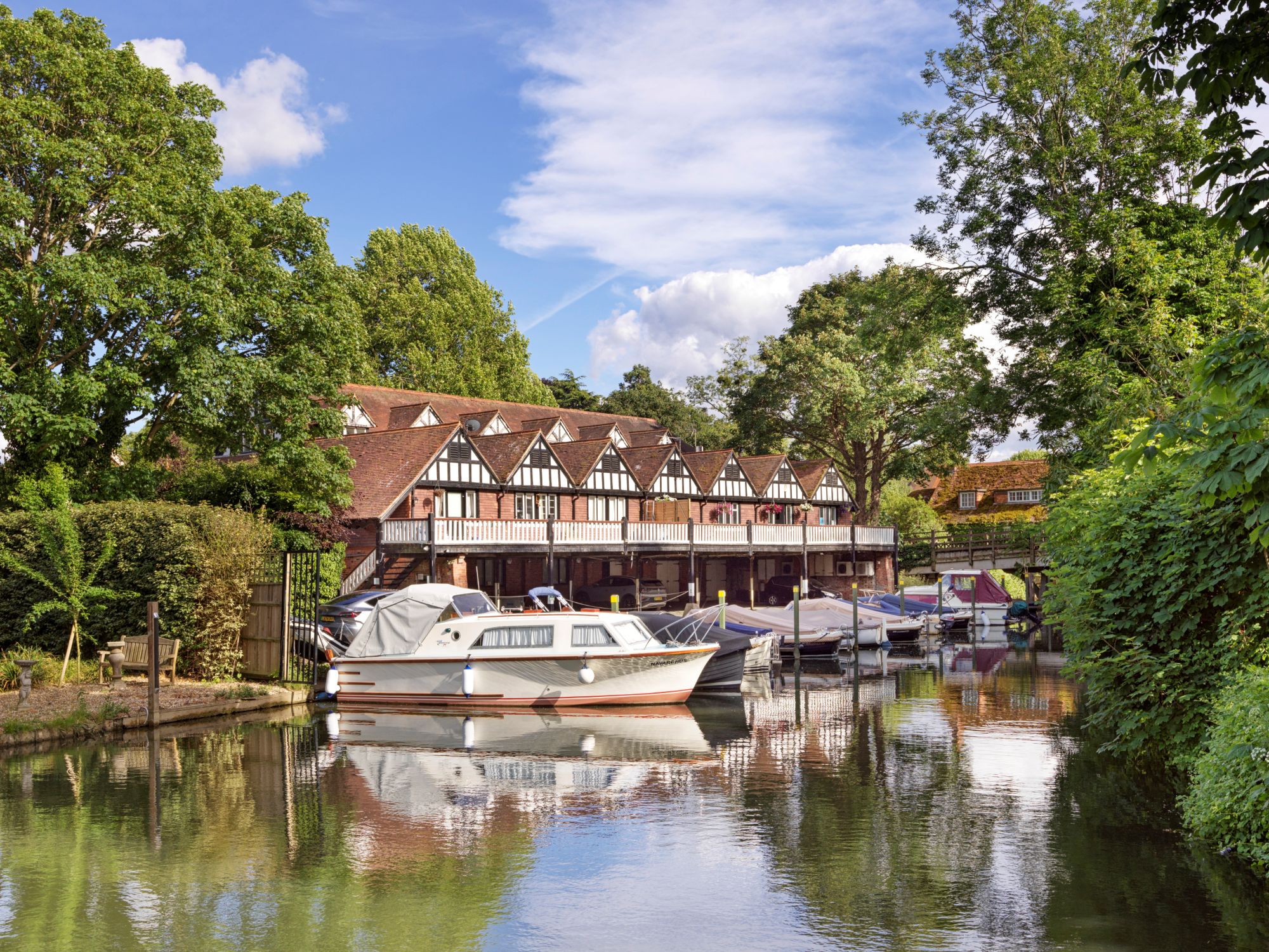 A photograph of a 2 storey Victorian boathouse. The ground floor is partly open and faces the river with boats moored in front. There is a balcony with timber balustrade running the length of the building which has a gable roof.
