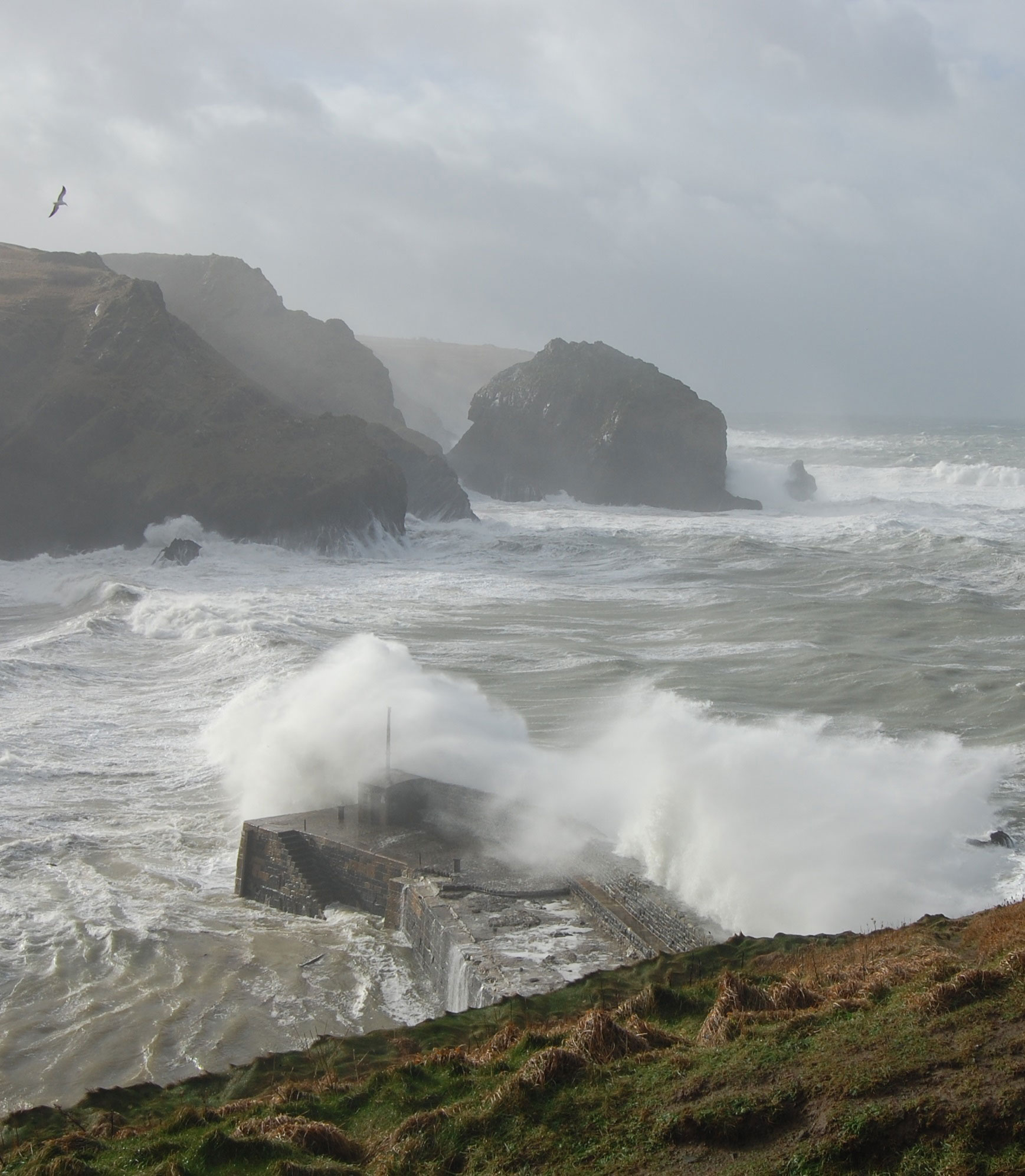 Photograph of a cove with waves crashing over a stone jetty and a rocky coastline in the background.