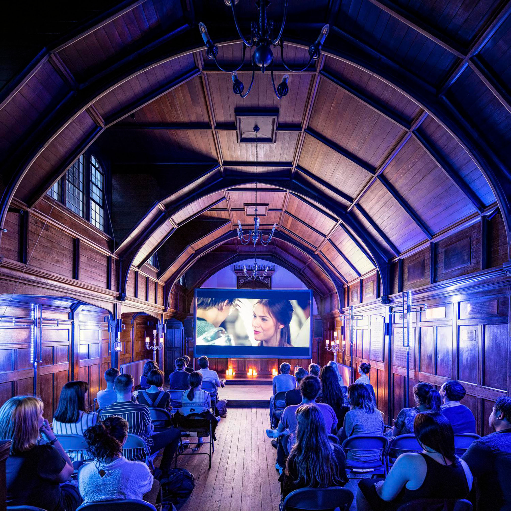 An audience watching a film in a wood-panelled hall with blue lighting and a large screen displaying a movie scene.