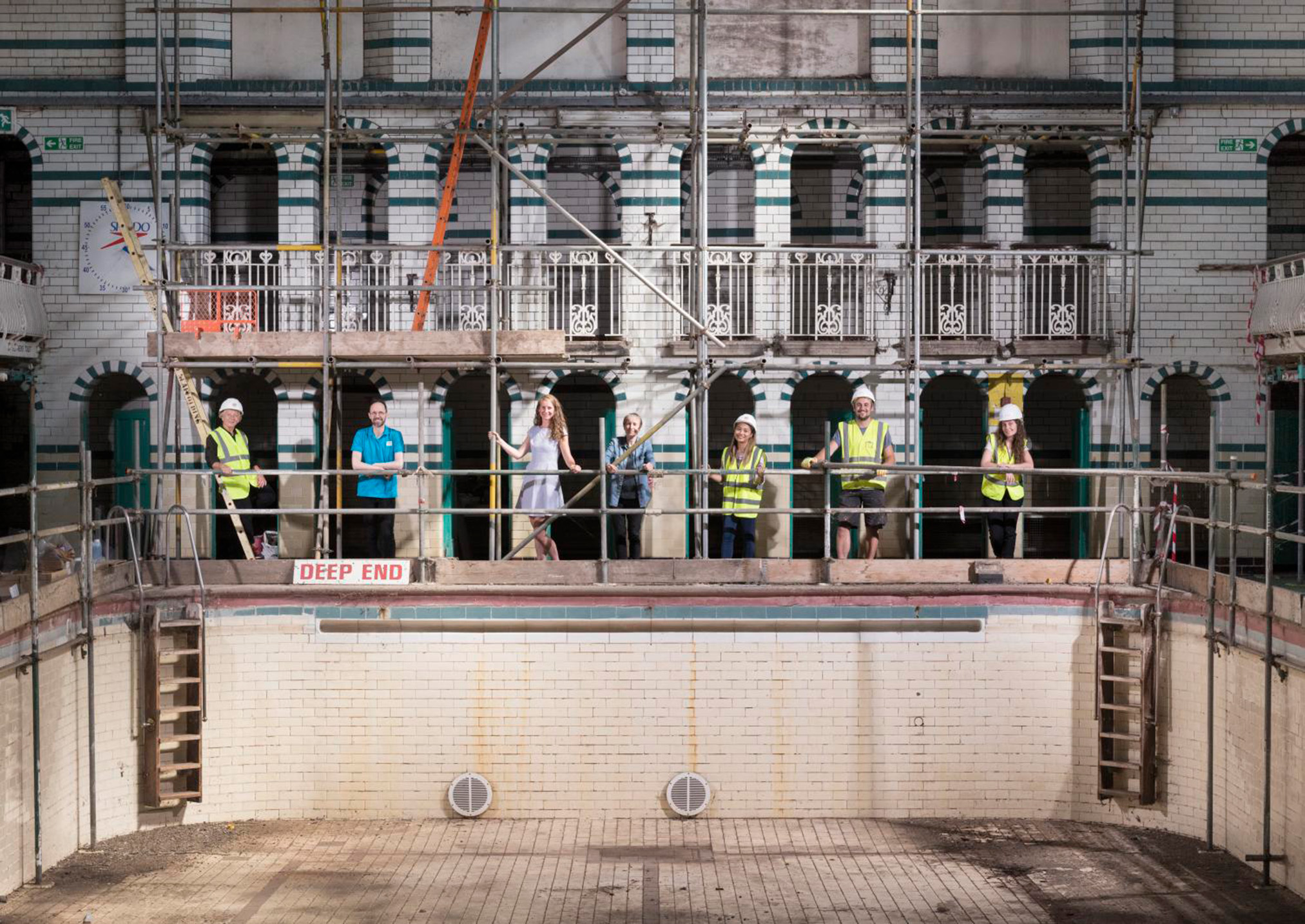 Seven people, four of whom are wearing yellow hi vis tops and white hard hats are standing under scaffolding at the deep end of an empty swimming pool. The interior of the building is tiled in white and dark green and the wall behind the people features twenty arches and decorative railing on the storey above the people.
