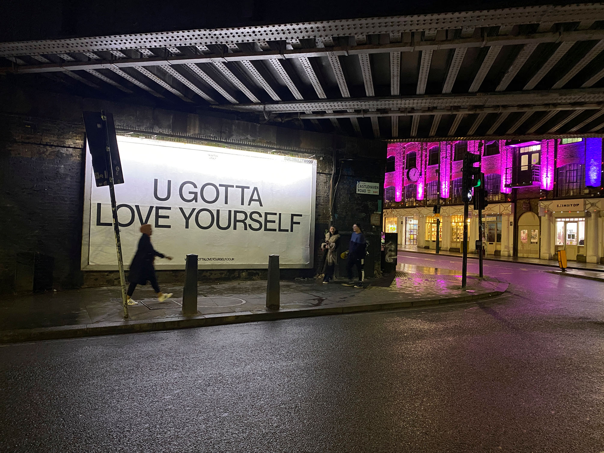 Pedestrians passing an illuminated billboard reading 'U Gotta Love Yourself' beneath the railway bridge on Castlehaven Road at night.