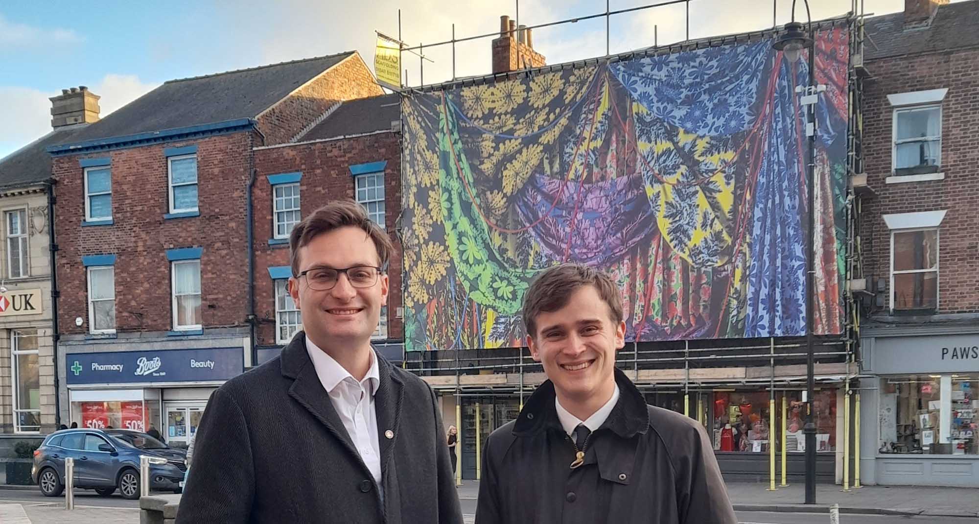 Two men standing next to each other in a high street. 