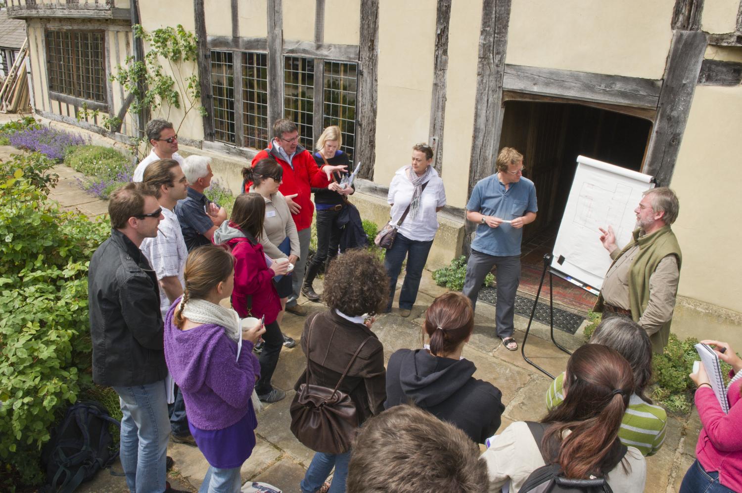 A group of people gathered outside a building, listening to a man speaking next to a flipchart. Some attendees are taking notes while others are observing.