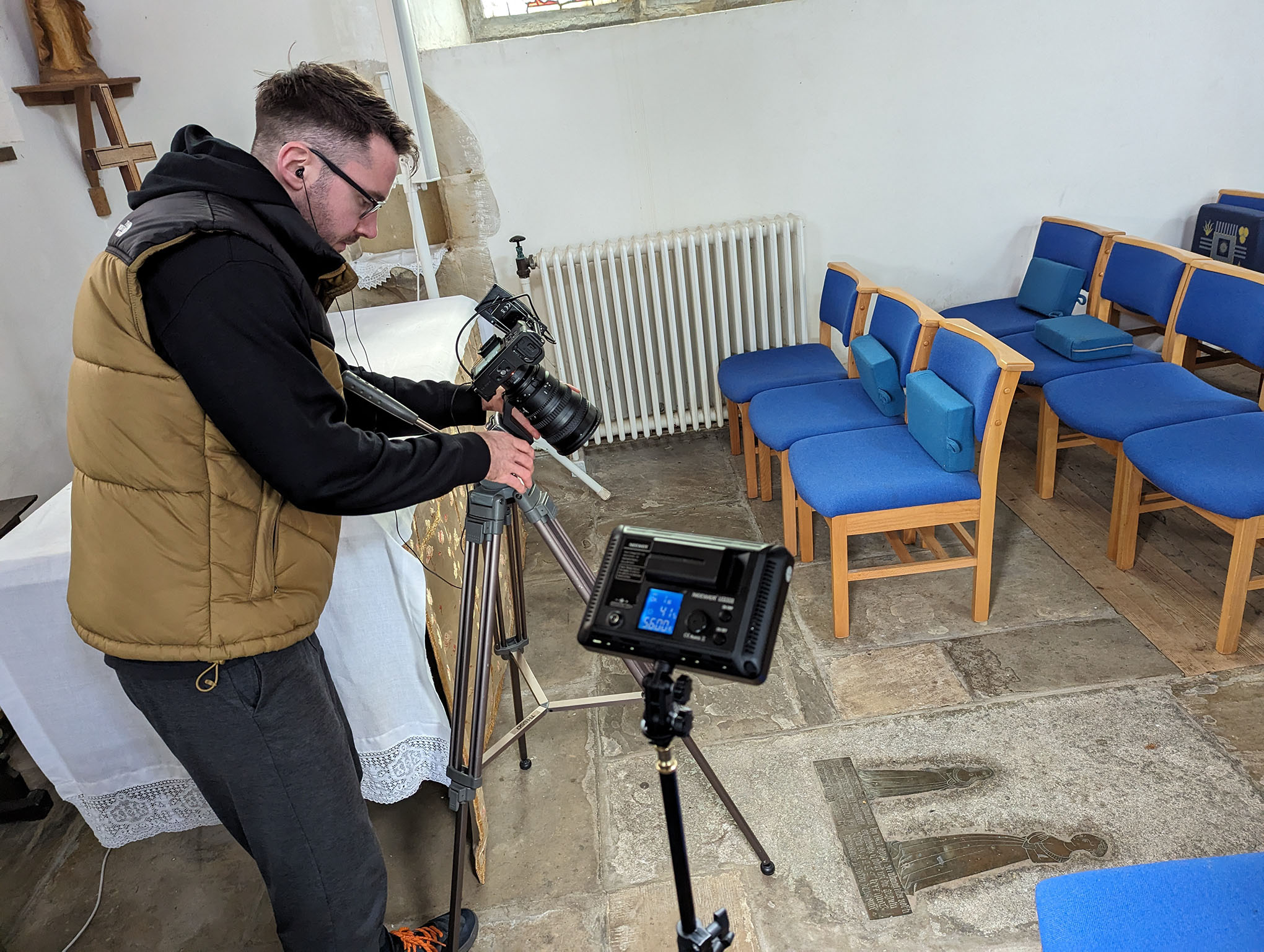 A man is stood next to a video camera on a tripod. He is adjusting it so it is pointing towards a brass plaque on the stone floor of a church. Chairs and an altar are visible behind the man and the camera. There is also a light on a tripod which is illuminating the brass plaque.