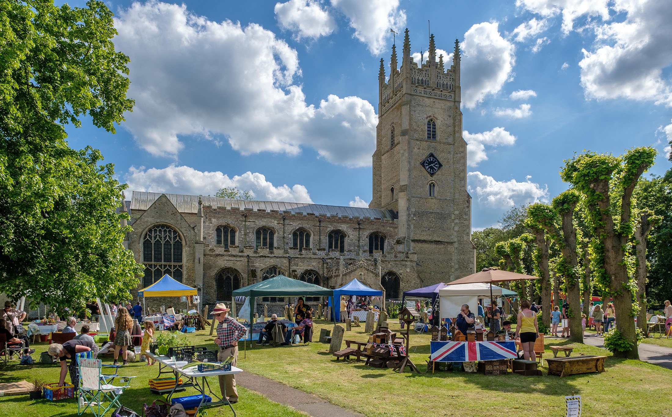 the photo shows the church on a sunny day with blue sky. On the lawn in front of the church are stalls and stands for a summer fair