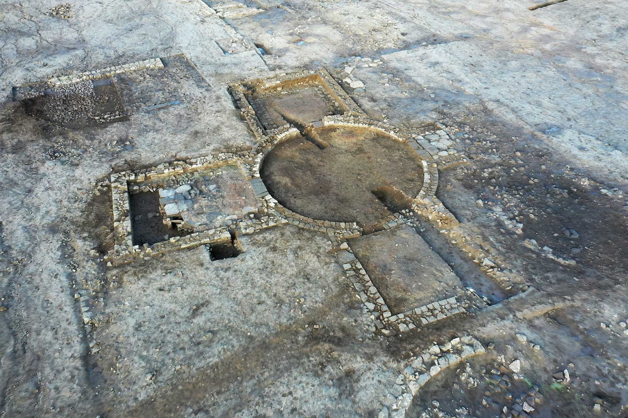 Low stone structures in excavated ground. The main structure includes a central circle of stone with three rectangular rooms radiating from it.