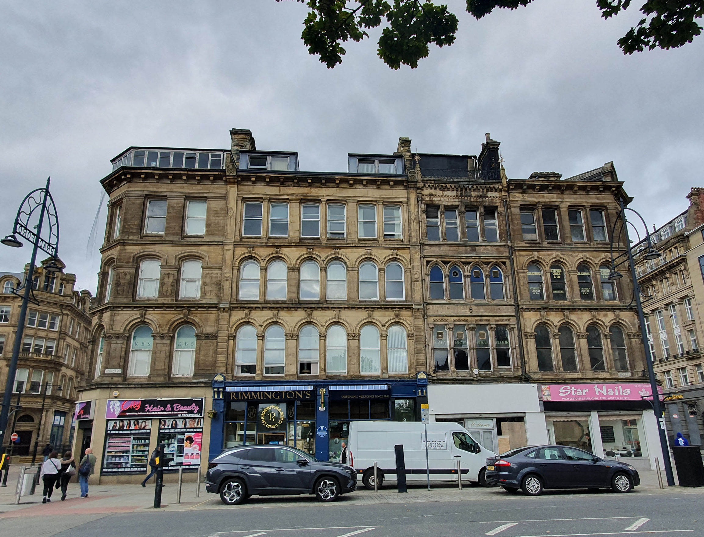 A photo of a street scene featuring cars, people and a 4-storey cream stone building with shop fronts on the ground floor.