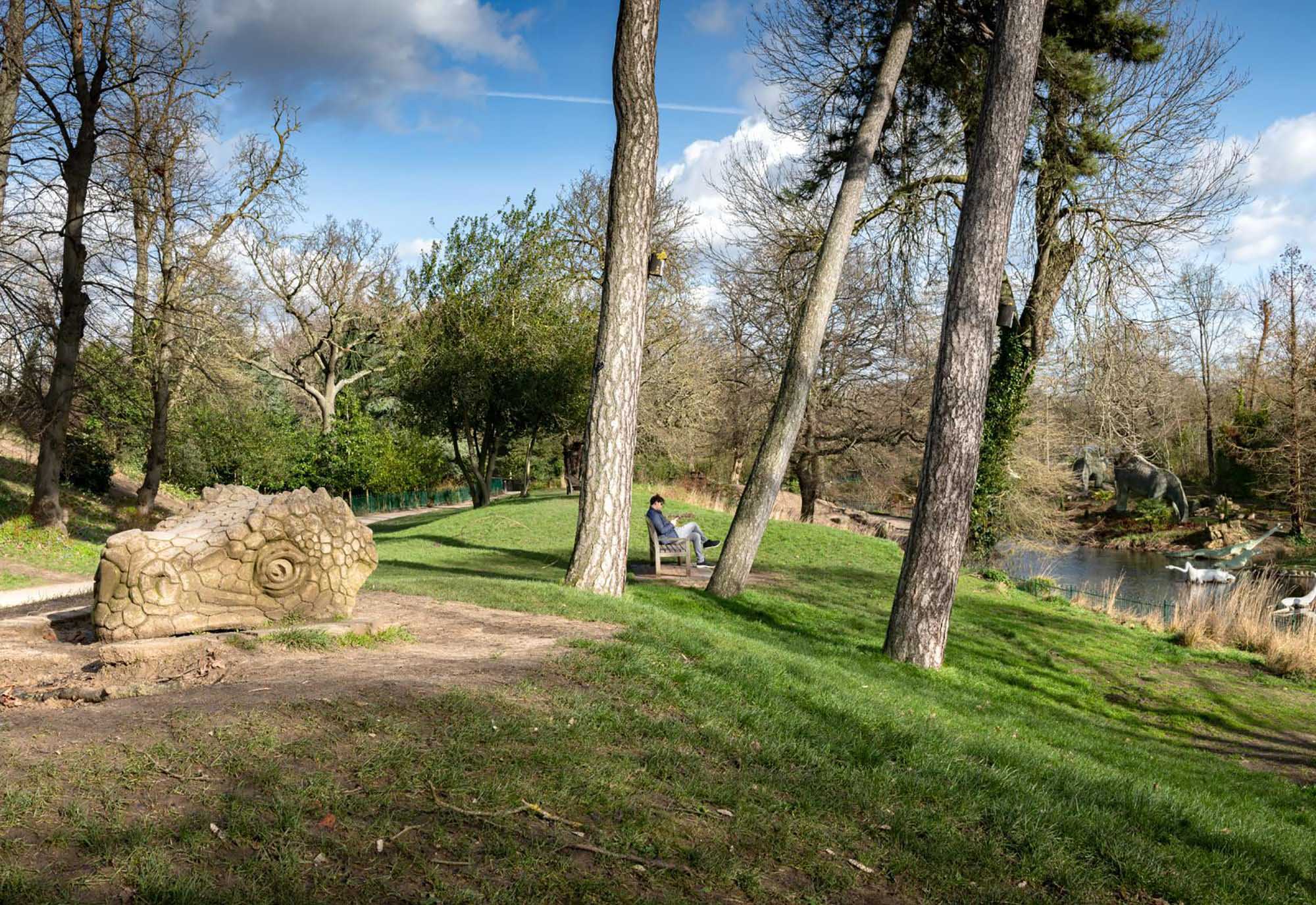 A man sits on a bench in parkland with dinosaur sculptures in front and behind him.