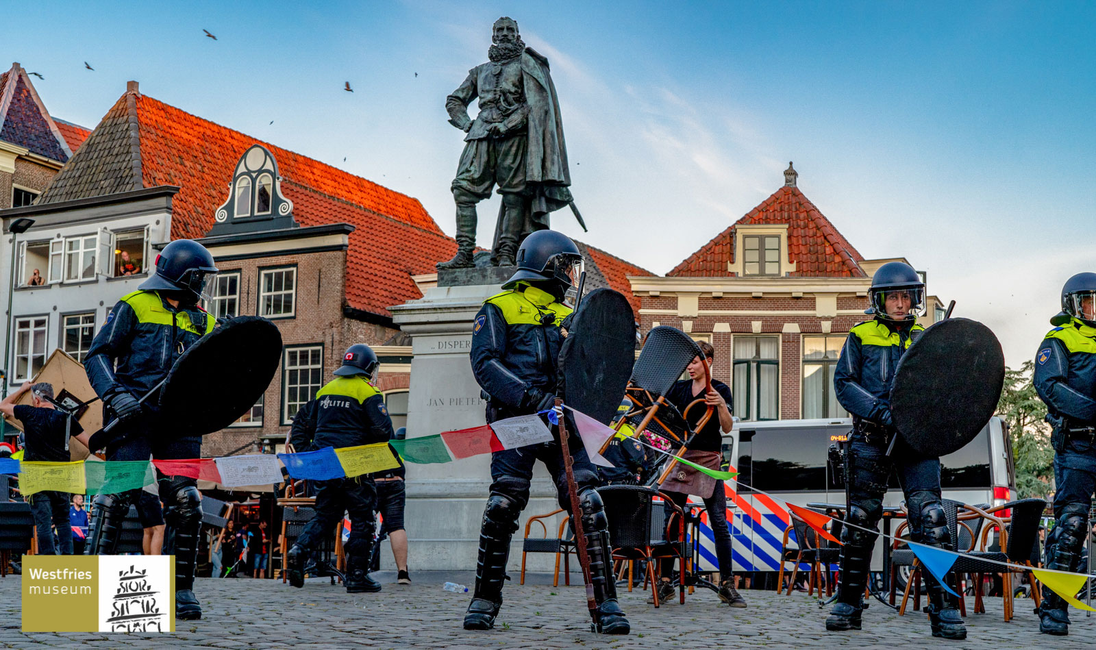 Dutch police in riot gear at a demonstration by a statue in a public space, with dutch town buildings in the background.