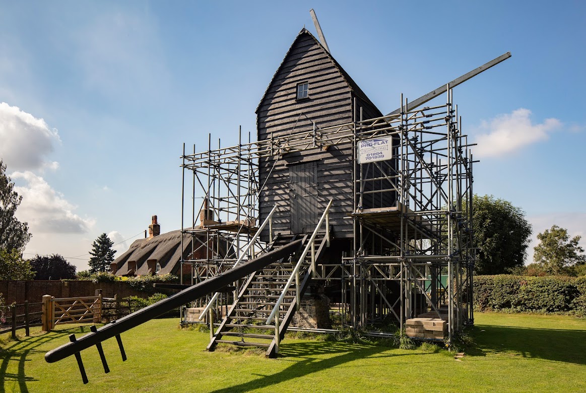 Bourn Windmill surrounded by scaffolding propping it  up. Steps are shown leading to a doorway. The post which turns the mill can be seen in the foreground. There's a small window towards the top of the mill and two sail posts can be seen on the other side of the building.