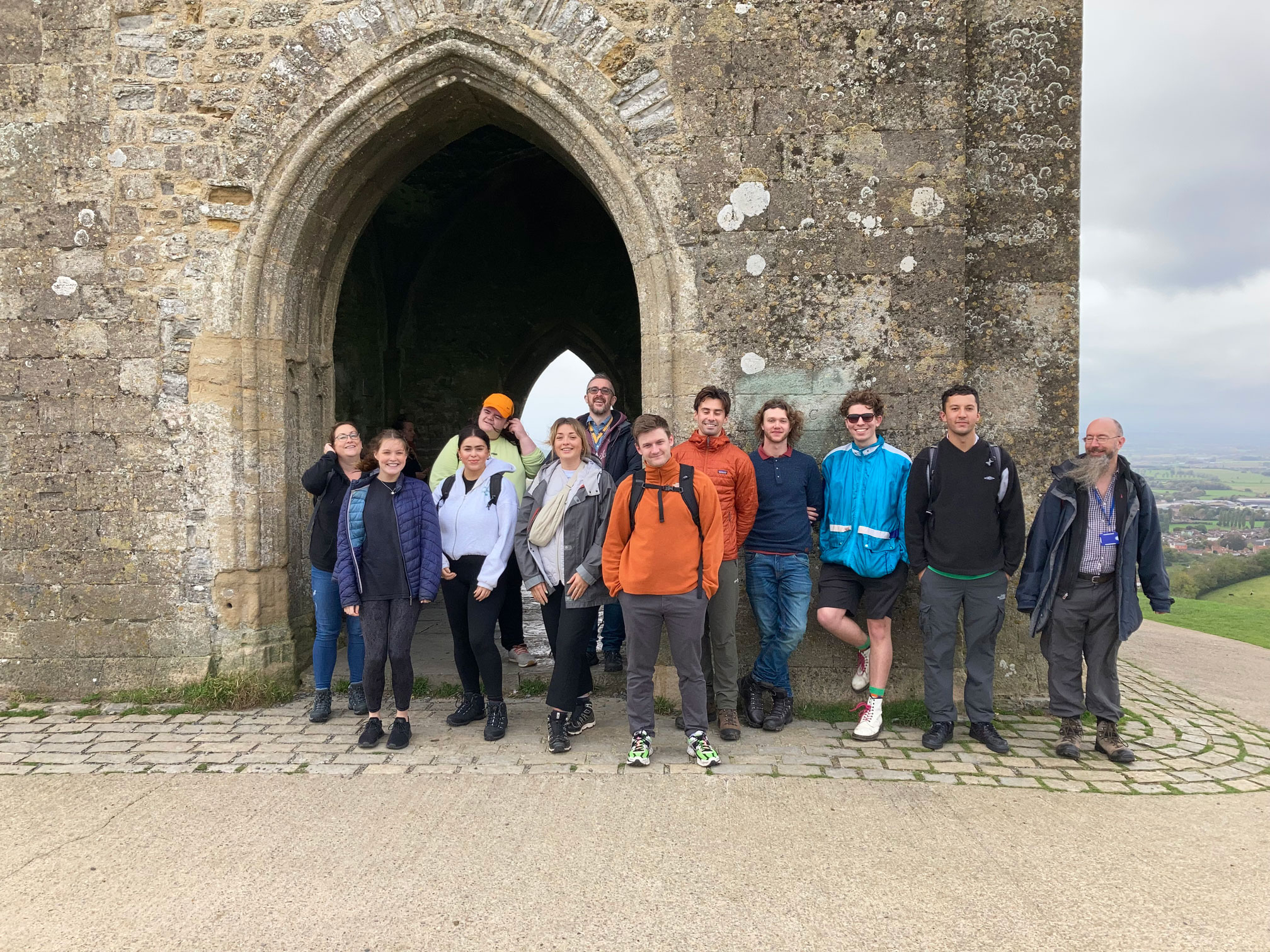 A group of smiling people posed in front of the archway of a church atop a tor.