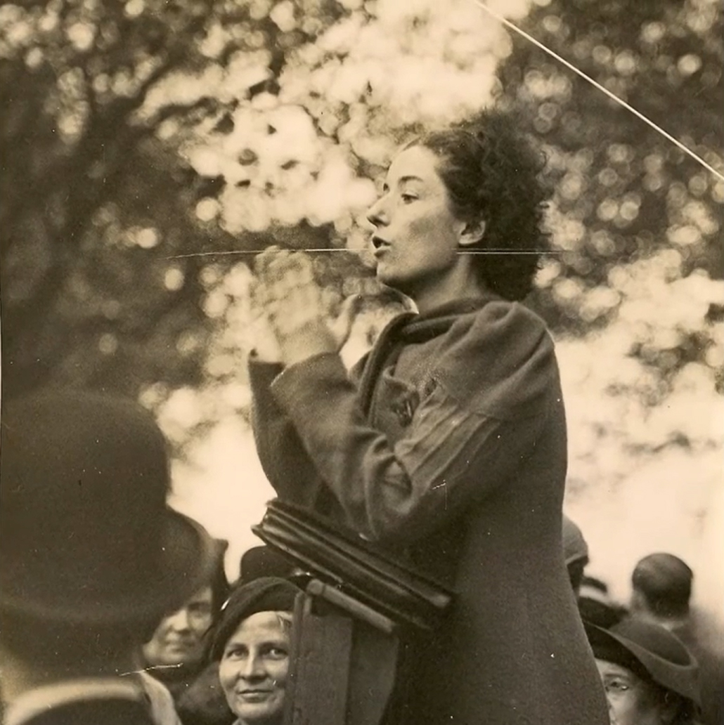 Black and white photo of a woman in profile, talking to a crowd of people from a rostrum.