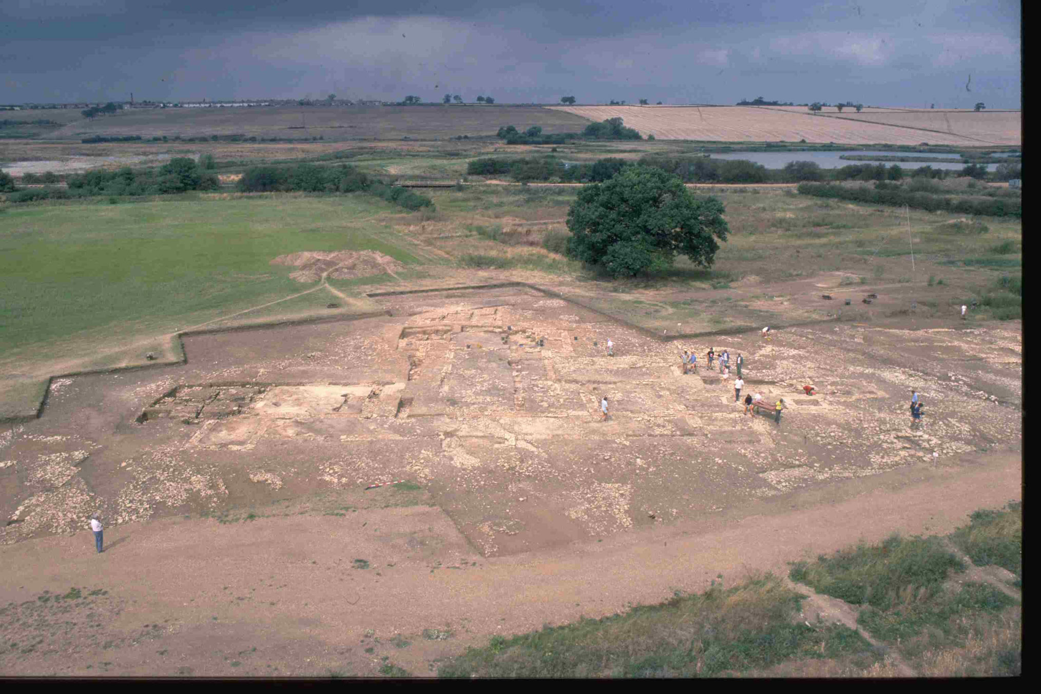 The foreground shows a large area of archaeological excavation with archaeologists just visible as small figures within the trench. Beyond the trench farmland stretches towards the horizon.