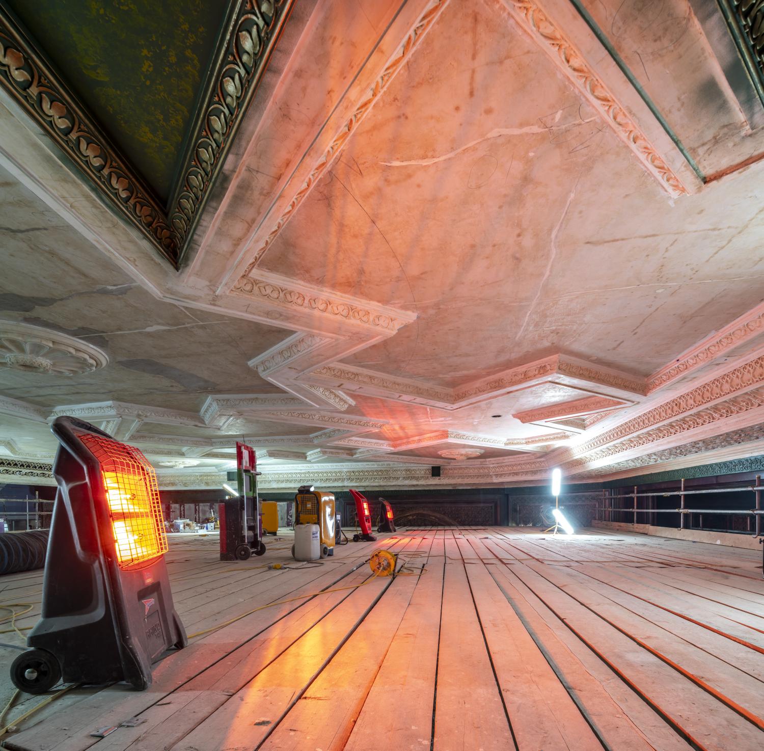 An electric heater rests on scaffolding underneath an ornate plaster ceiling, with other heaters and dehumidifiers in the background.