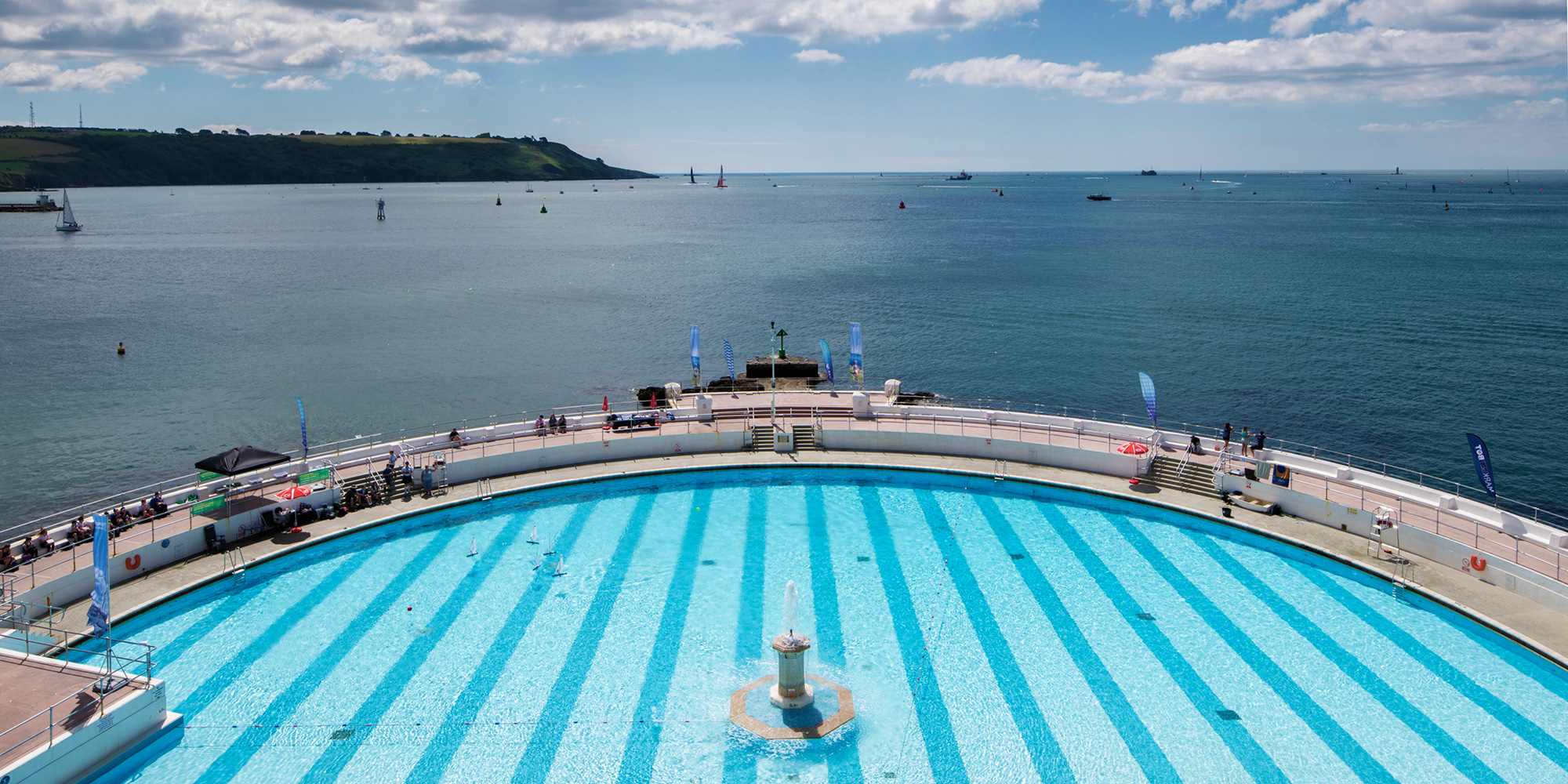 A stiped-blue-bottomed open-air swimming pool looks out over a seascape, with model sailing ships in the pool echoing the sails in the bay.