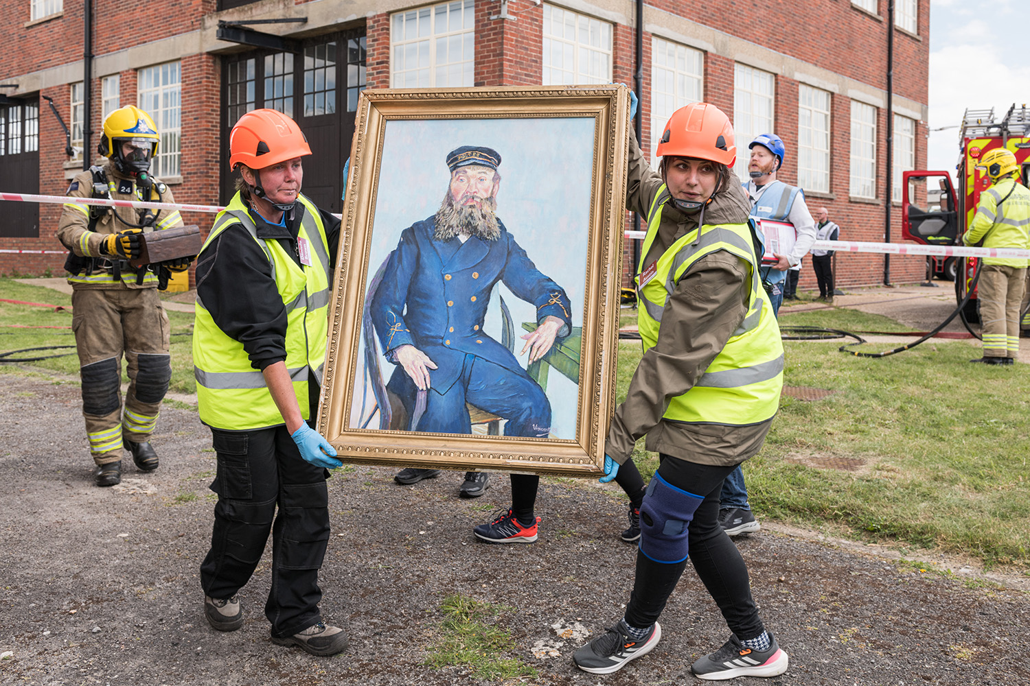 Two people dressed in protective clothing carry a large painting of a sailor between them.