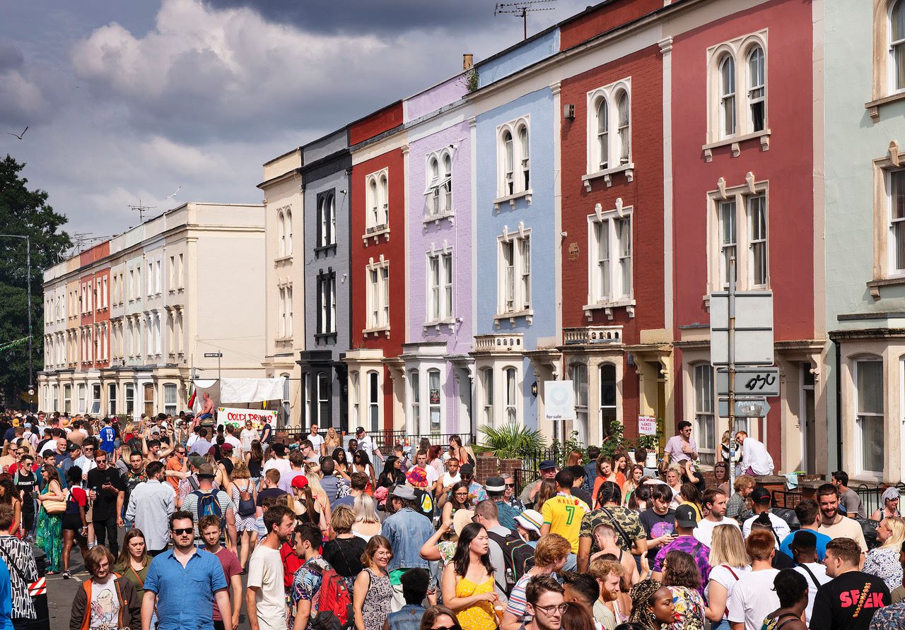 A street scene full of people in the foreground and coloured townhouses in the background.